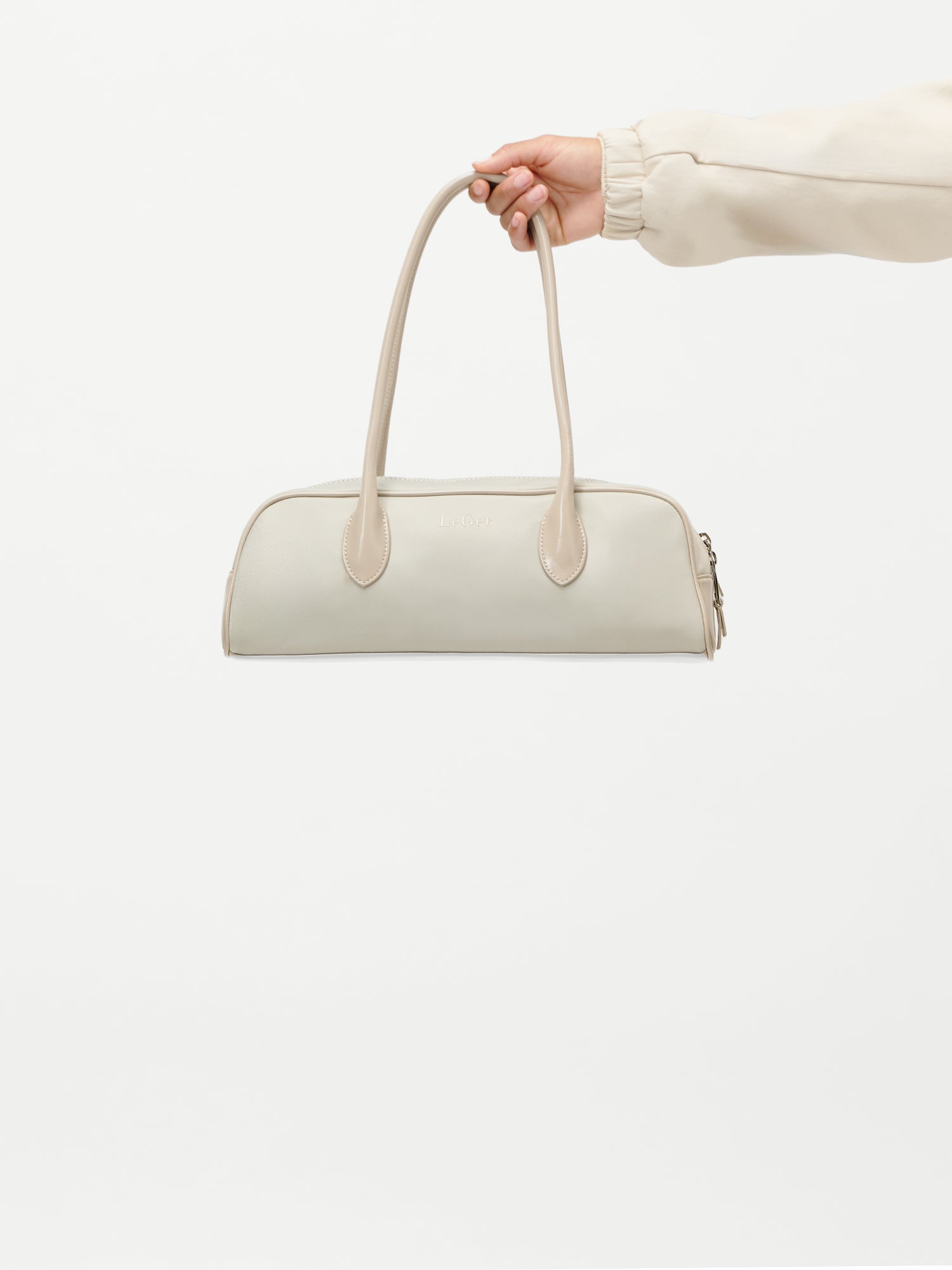 Eine Hand hält die LeGer by Lena Gercke Frauke Bag, eine beigefarbene rechteckige Handtasche mit zwei langen Henkeln, vor einem schlichten weißen Hintergrund. Die Person trägt ein helles, langärmeliges Oberteil.