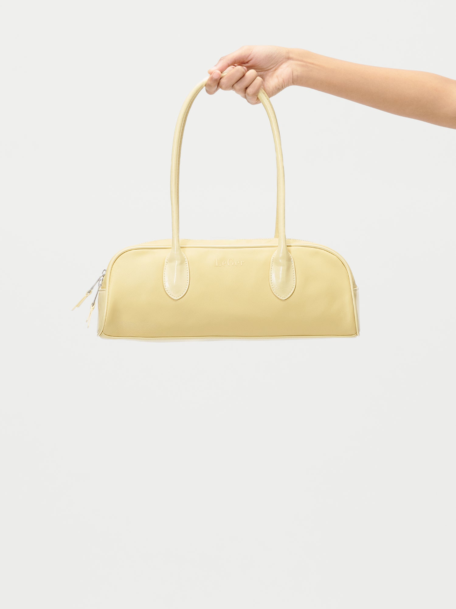 Eine Hand hält die Frauke Bag von LeGer by Lena Gercke hoch, eine hellgelbe Handtasche mit langen Henkeln und dezenten Nähten, die ein schlichtes rechteckiges Design auf weißem Grund zeigt.
