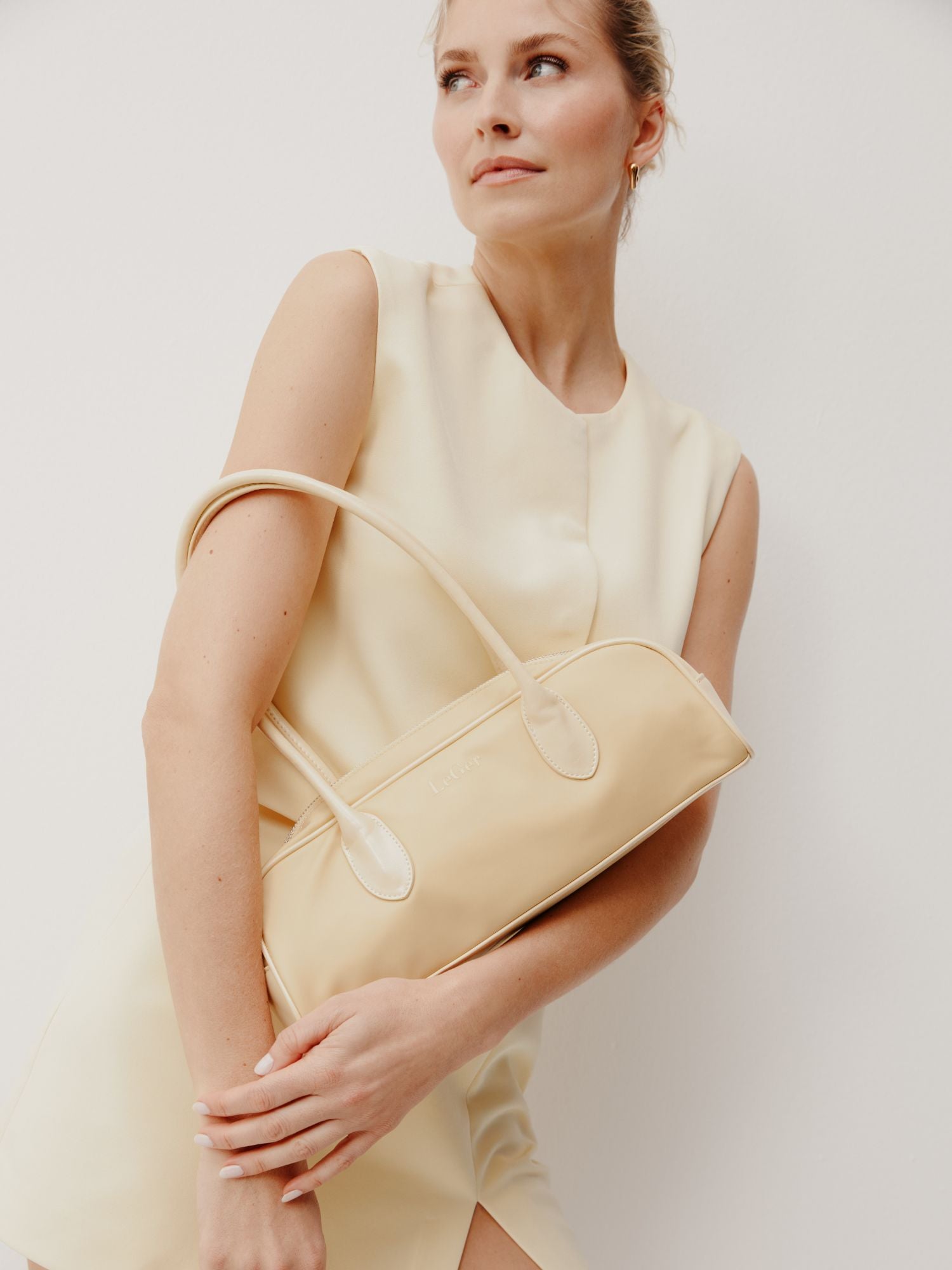 Eine Frau in einem ärmellosen cremefarbenen Kleid posiert vor einem schlichten weißen Hintergrund. Sie hält die LeGer by Lena Gercke Frauke Bag, eine beigefarbene Handtasche, unter ihrem Arm und blickt mit einem sanften Ausdruck leicht nach oben.