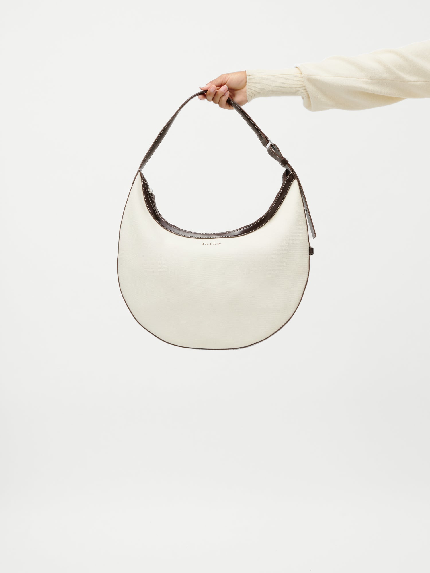 Eine Hand in einem cremefarbenen Ärmel hält die Tasche „Gisa“ aus der LeGer by Lena Gercke-Kollektion – ein weißes, halbmondförmiges Accessoire mit dunkelbrauner Einfassung und einem kurzen Riemen –, die vor einem schlichten weißen Hintergrund zu sehen ist.