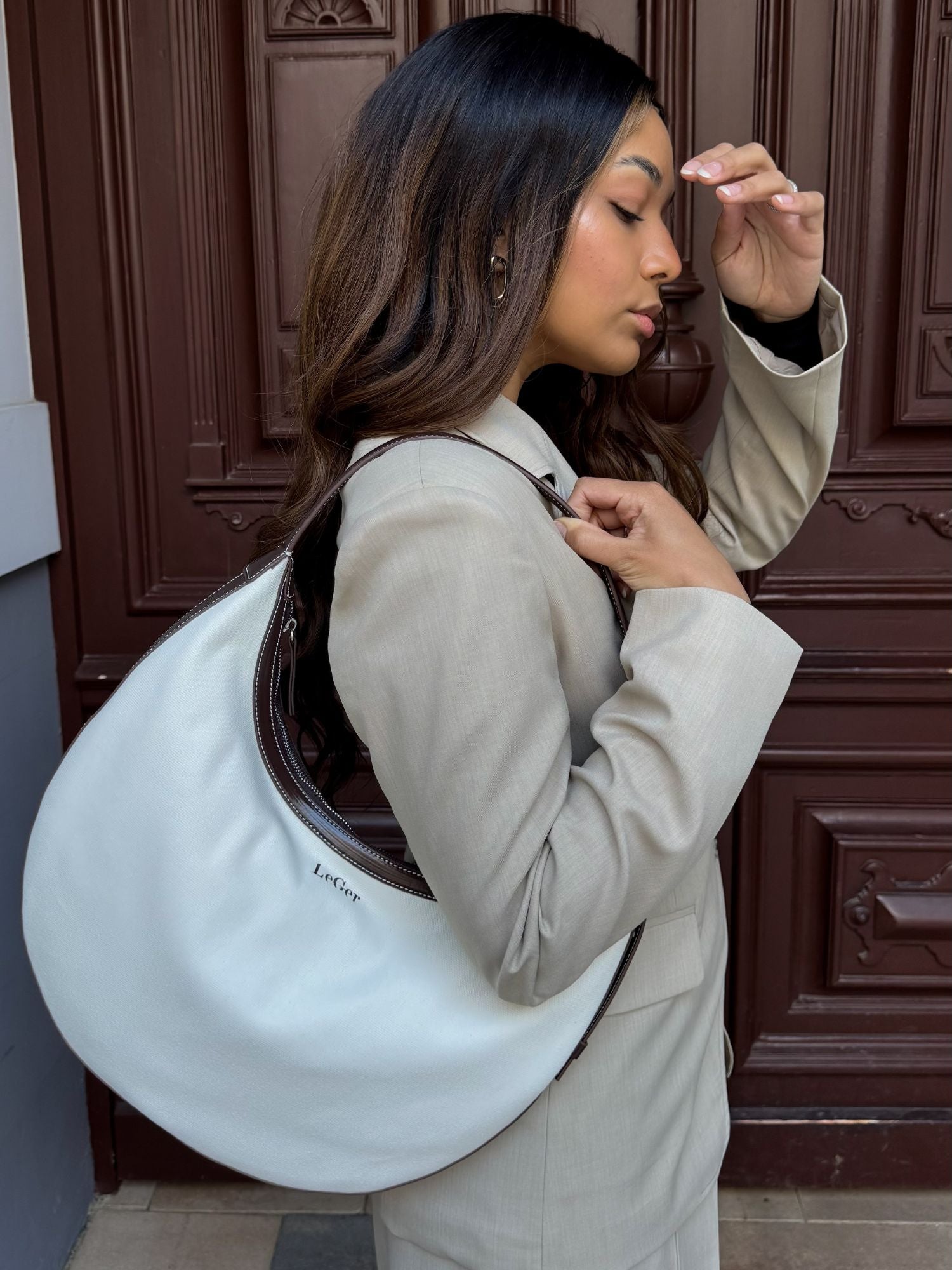 Eine Frau mit langen braunen Haaren in einem hellbeigen Blazer trägt die große Gisa Bag von LeGer by Lena Gercke in Halbmond-Form über der Schulter und steht vor einer dunklen Holztür, um einen stilvollen Kontrast zu schaffen, während sie sich an die Stirn fasst.