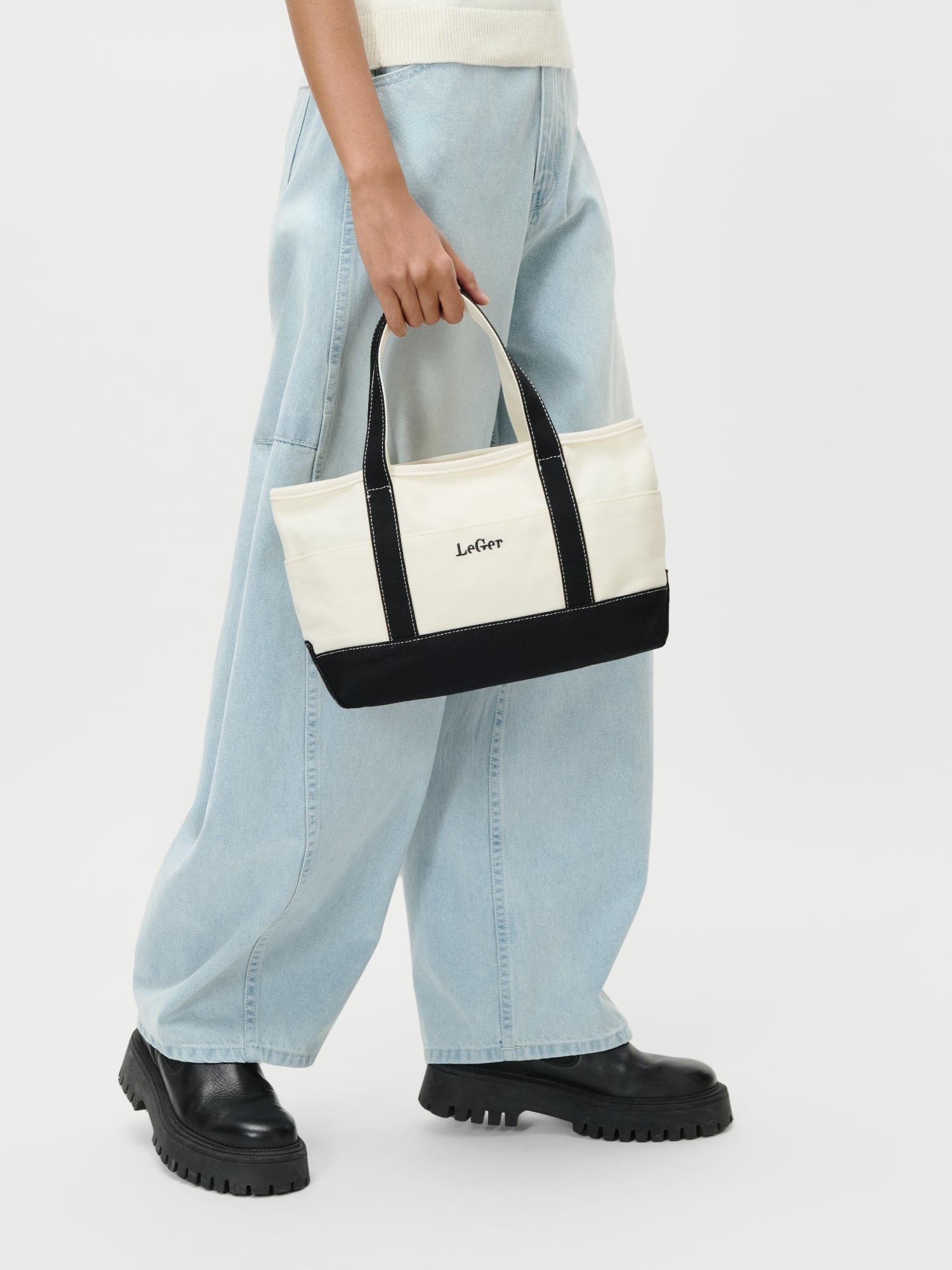 Eine Person in hellblauen, weit geschnittenen Jeans und schwarzen, klobigen Stiefeln hält eine weiße und schwarze Sheila Bag von LeGer by Lena Gercke vor einem schlichten weißen Hintergrund.
