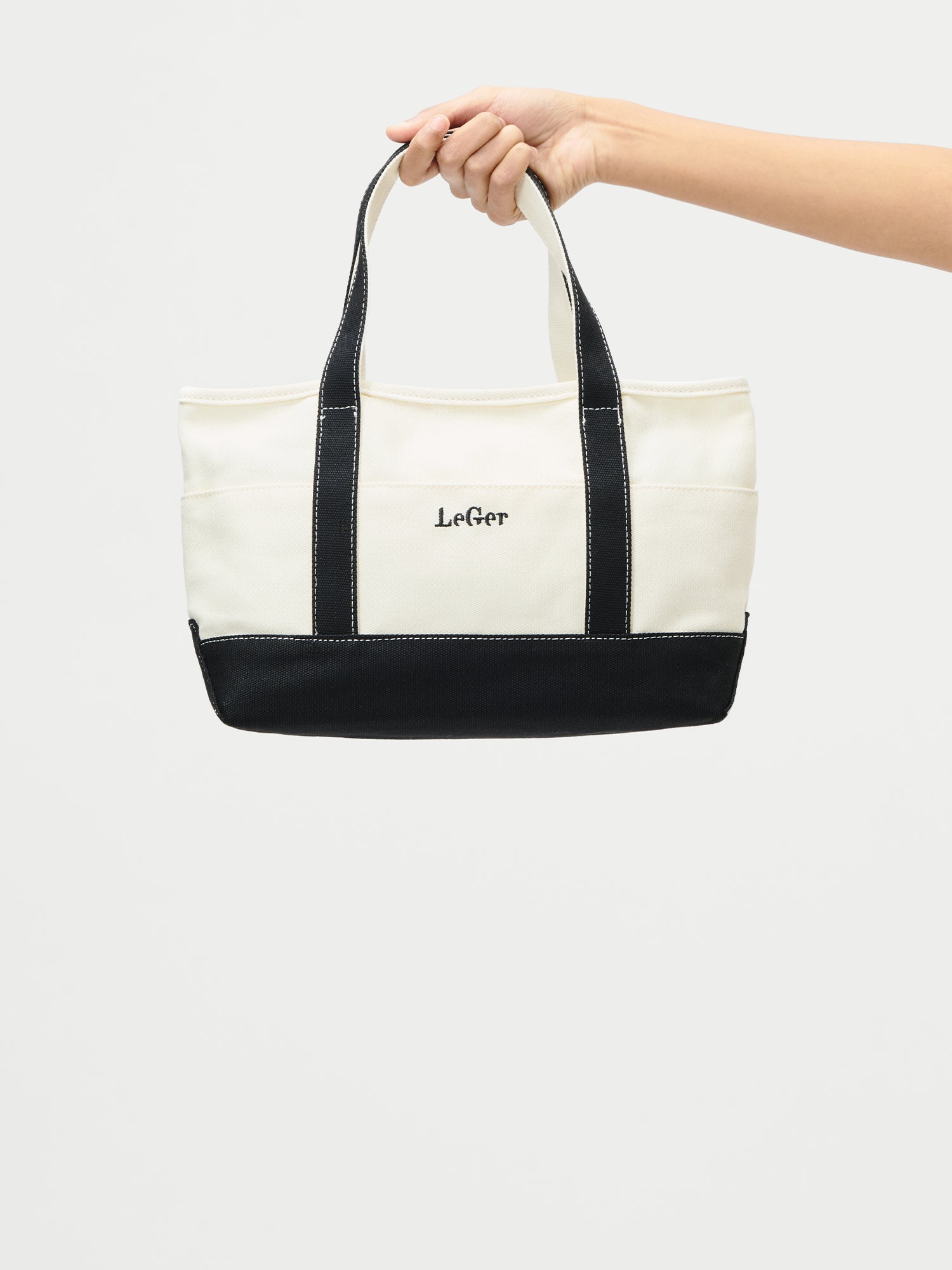 Eine Hand hält die Sheila Bag von LeGer by Lena Gercke, eine zweifarbige Canvas-Tasche mit schwarzen Griffen und Boden und einem weißen Korpus mit dem Markennamen auf der Vorderseite.