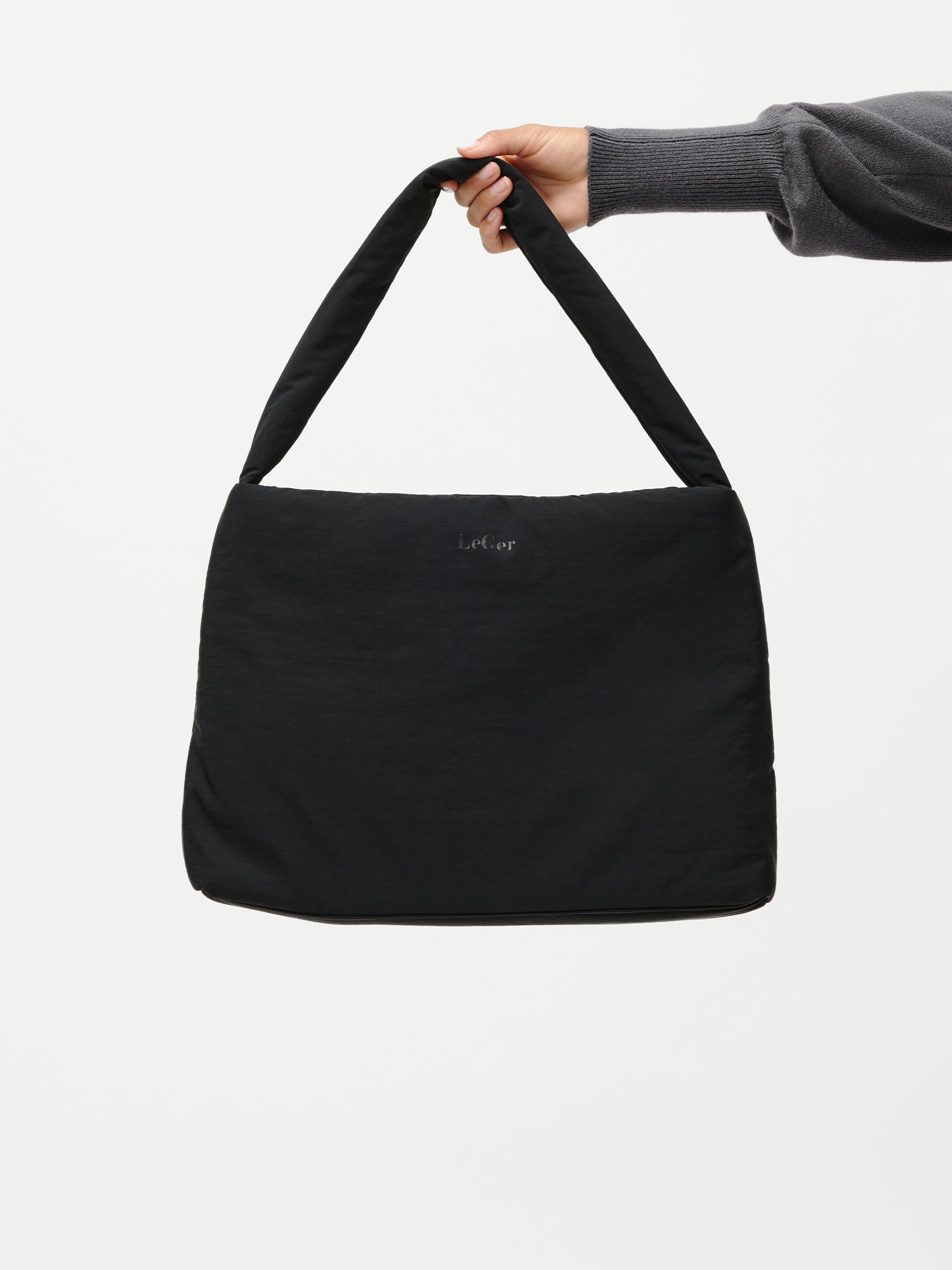Eine Person in einem dunklen, langärmeligen Oberteil hält die Katrin Bag von LeGer by Lena Gercke - eine schlichte schwarze Umhängetasche mit einem breiten Riemen - vor einem weißen Hintergrund.