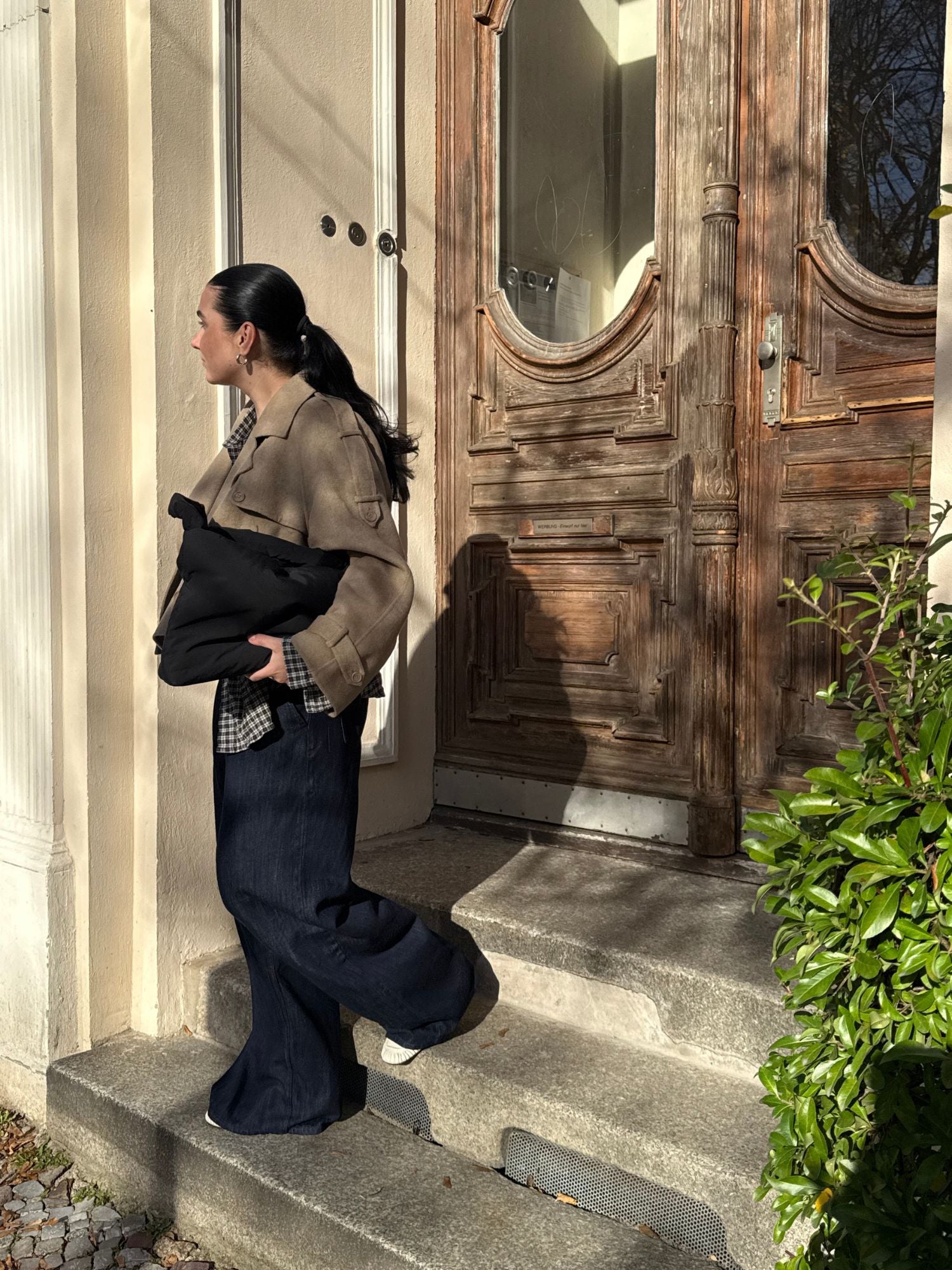 Eine Frau steht im Sonnenlicht auf einer Steintreppe. Sie trägt eine beige Jacke und eine Hose mit weitem Bein sowie die Tasche „Katrin“ von LeGer by Lena Gercke. Sie posiert vor Holztüren mit Glaseinsätzen; die Sträucher in der Nähe unterstreichen das minimalistische Design.