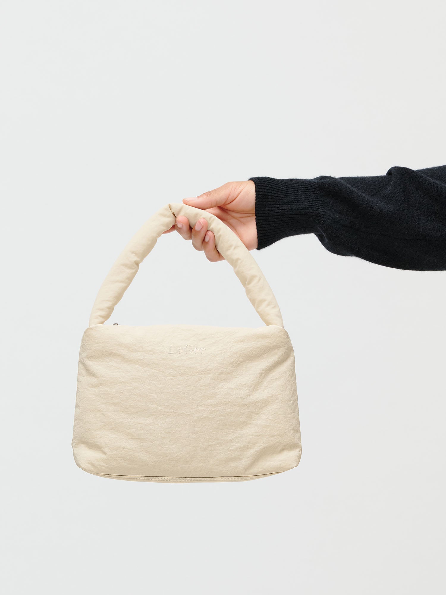 Eine Person, die ein schwarzes langärmeliges Oberteil trägt, hält die Lucky Bag von LeGer by Lena Gercke, eine kleine rechteckige beigefarbene Handtasche mit einem gepolsterten Henkel, vor einem schlichten weißen Hintergrund.