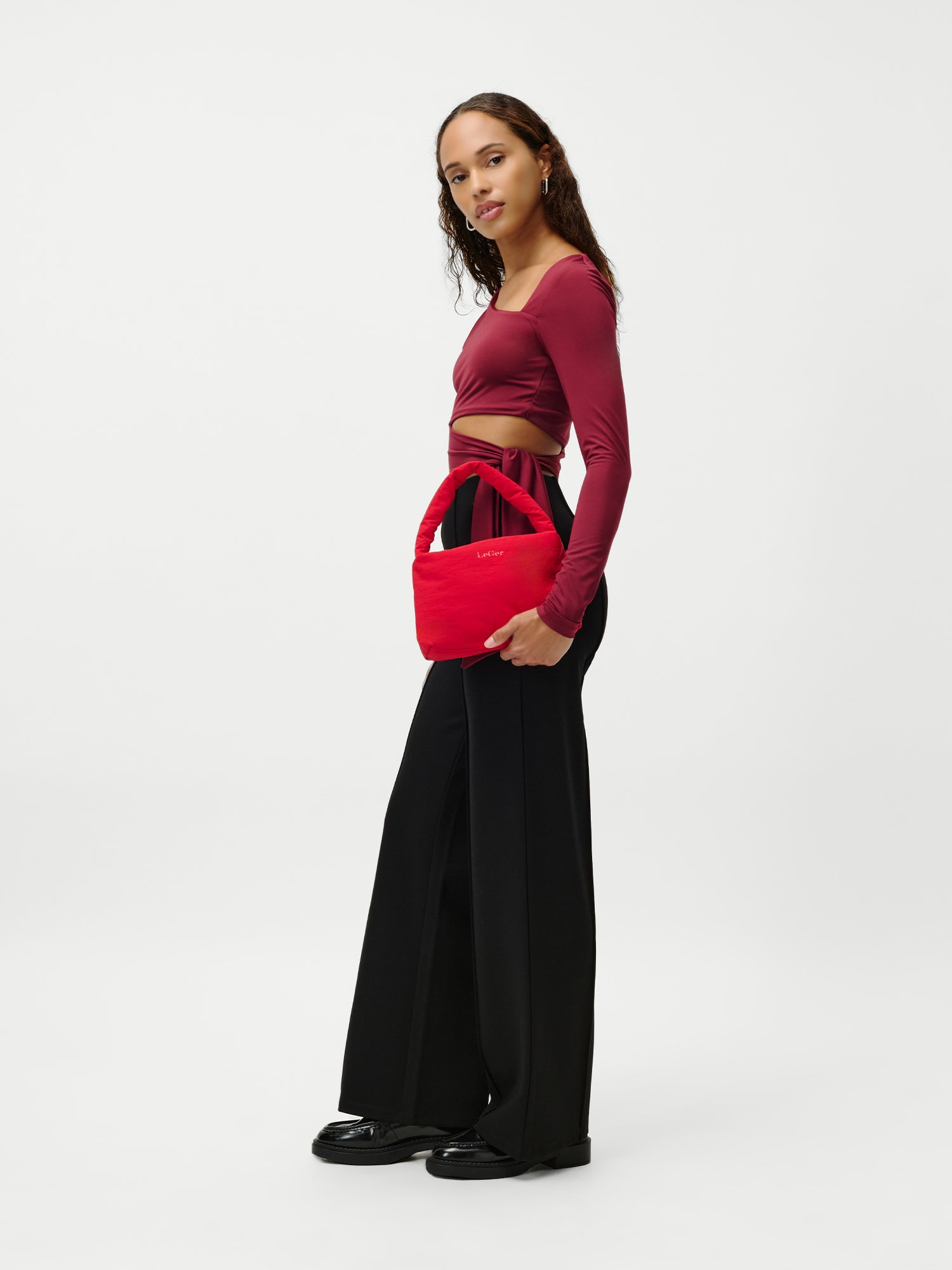 Eine Frau steht vor einem schlichten weißen Hintergrund, trägt ein kastanienbraunes, langärmeliges Crop-Top, eine schwarze, hochgeschlossene Hose mit weitem Bein, schwarze Schuhe und hält die LeGer by Lena Gercke Lucky Bag, eine kleine, leuchtend rote Handtasche.