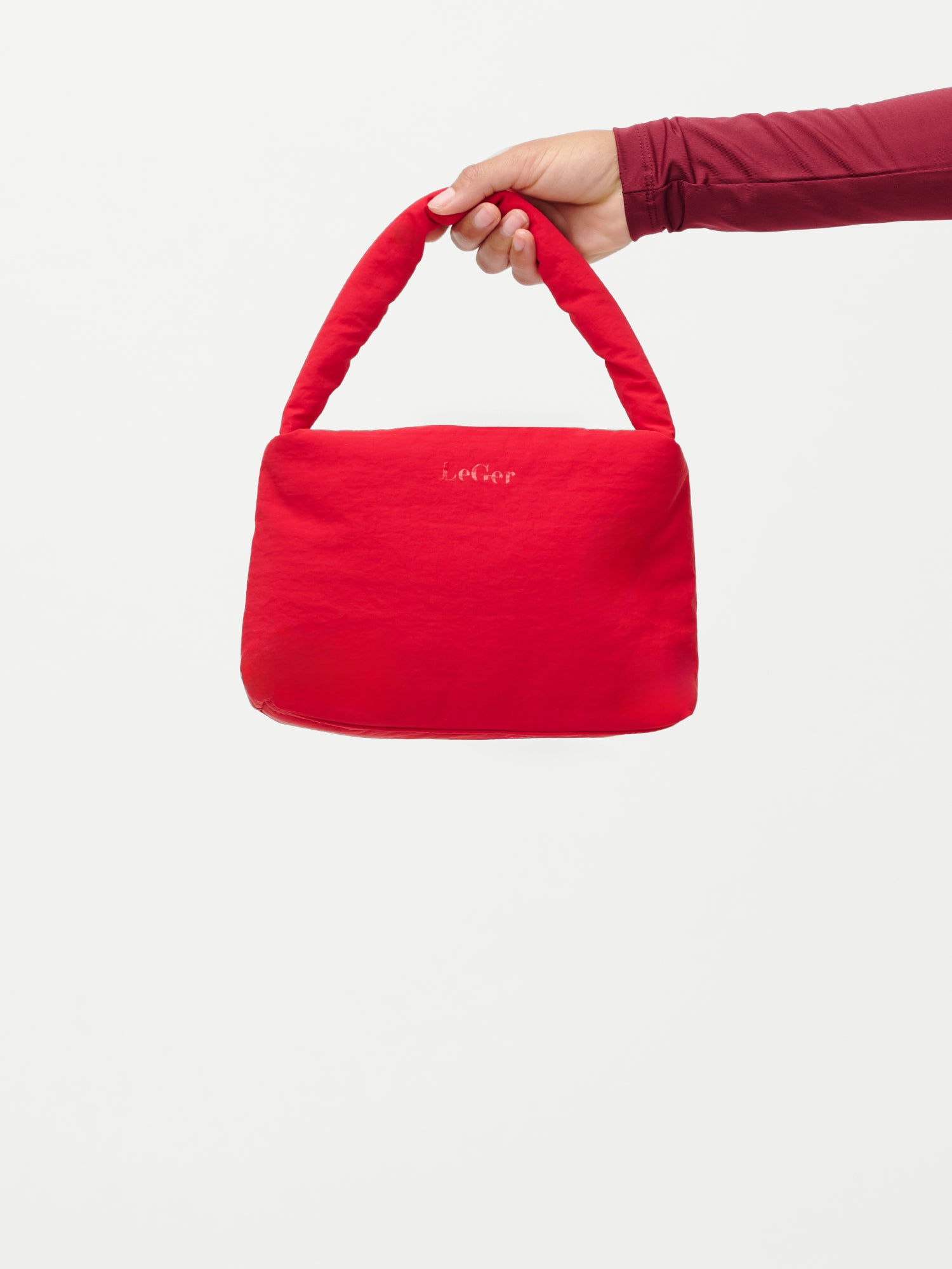 Eine Hand in einem kastanienbraunen, langärmeligen Oberteil hält die kleine, leuchtend rote Lucky Bag von LeGer by Lena Gercke. Sie hat eine weiche, bauschige Textur, einen kurzen Henkel und den Aufdruck "Lecker" in Weiß auf einem schlichten weißen Hintergrund.