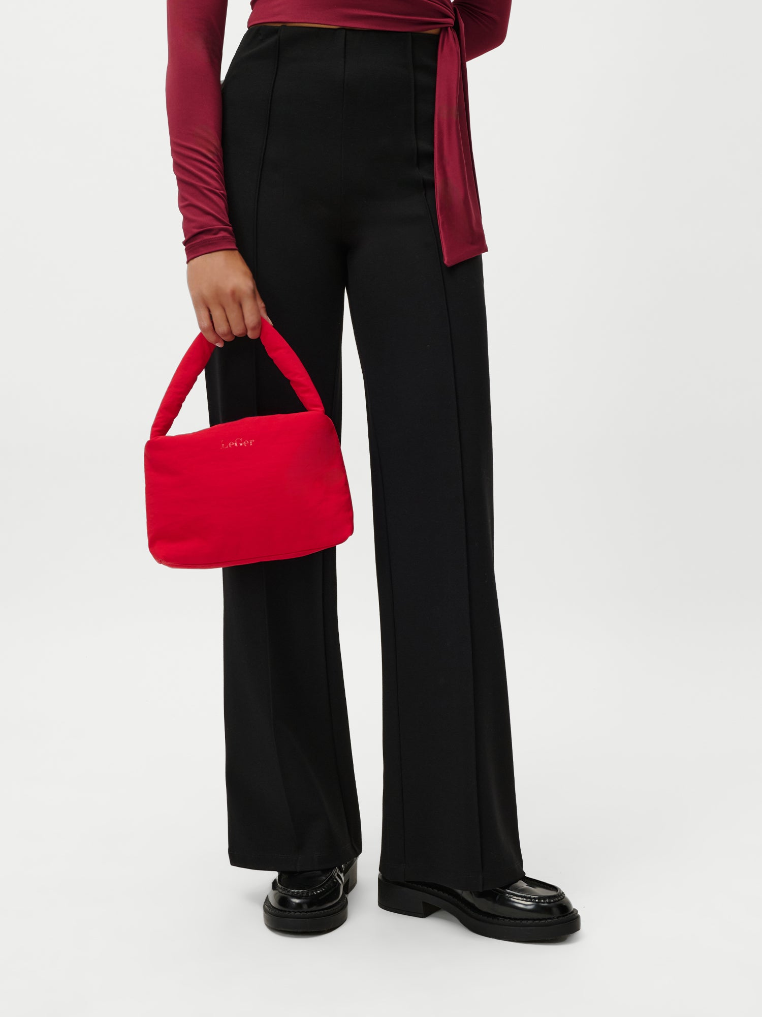 Eine Person in einer hochtaillierten schwarzen Hose, einem langärmeligen burgunderroten Oberteil und schwarzen Schuhen hält die LeGer by Lena Gercke Lucky Bag - eine kleine rote Handtasche - vor einem schlichten weißen Hintergrund.