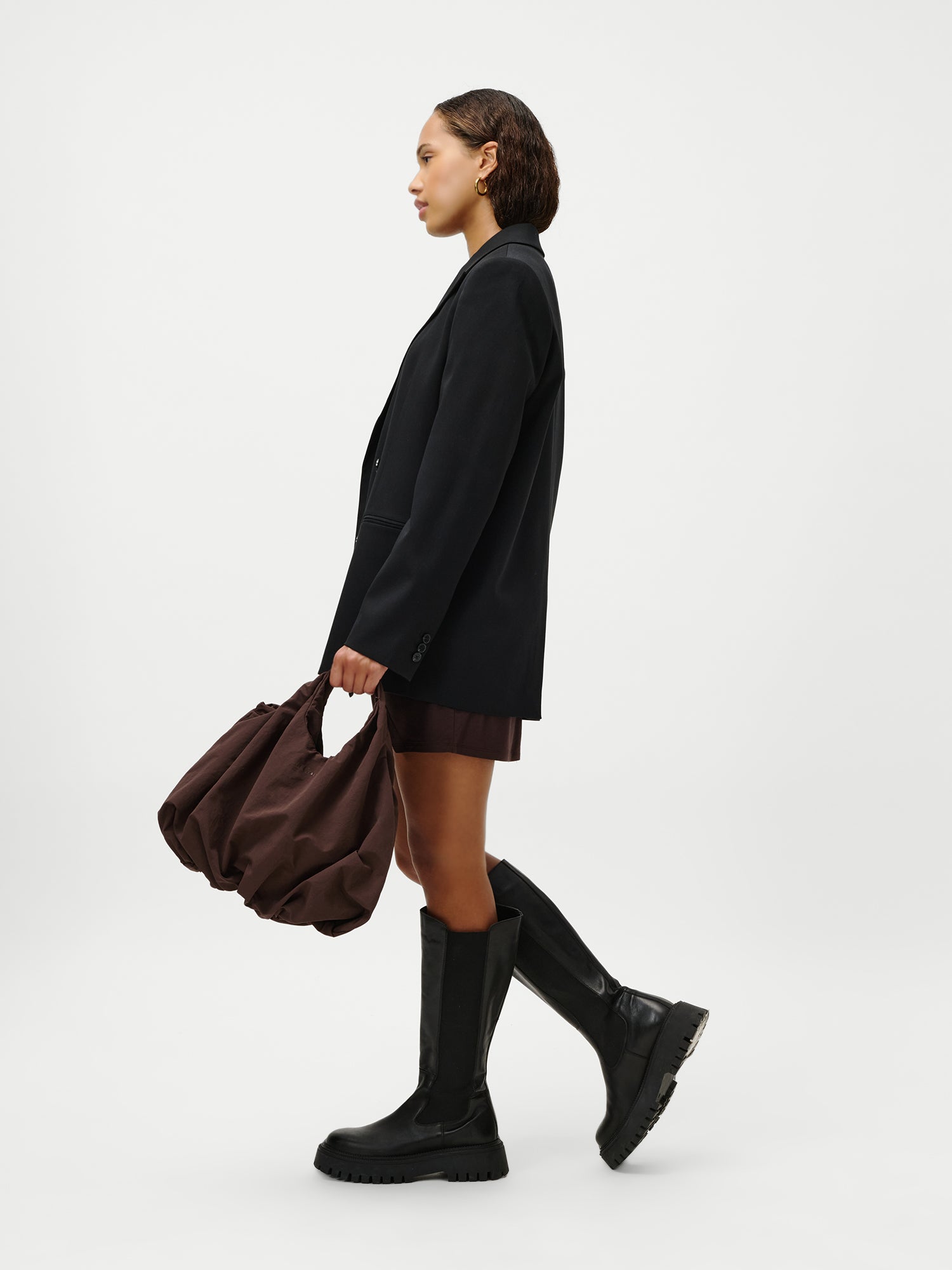 Eine Frau im Profil vor weißem Hintergrund trägt einen schwarzen Blazer, einen braunen Minirock, schwarze kniehohe Stiefel und trägt die LeGer by Lena Gercke Lotti Bag - eine große braune Tasche - in ihrer linken Hand.