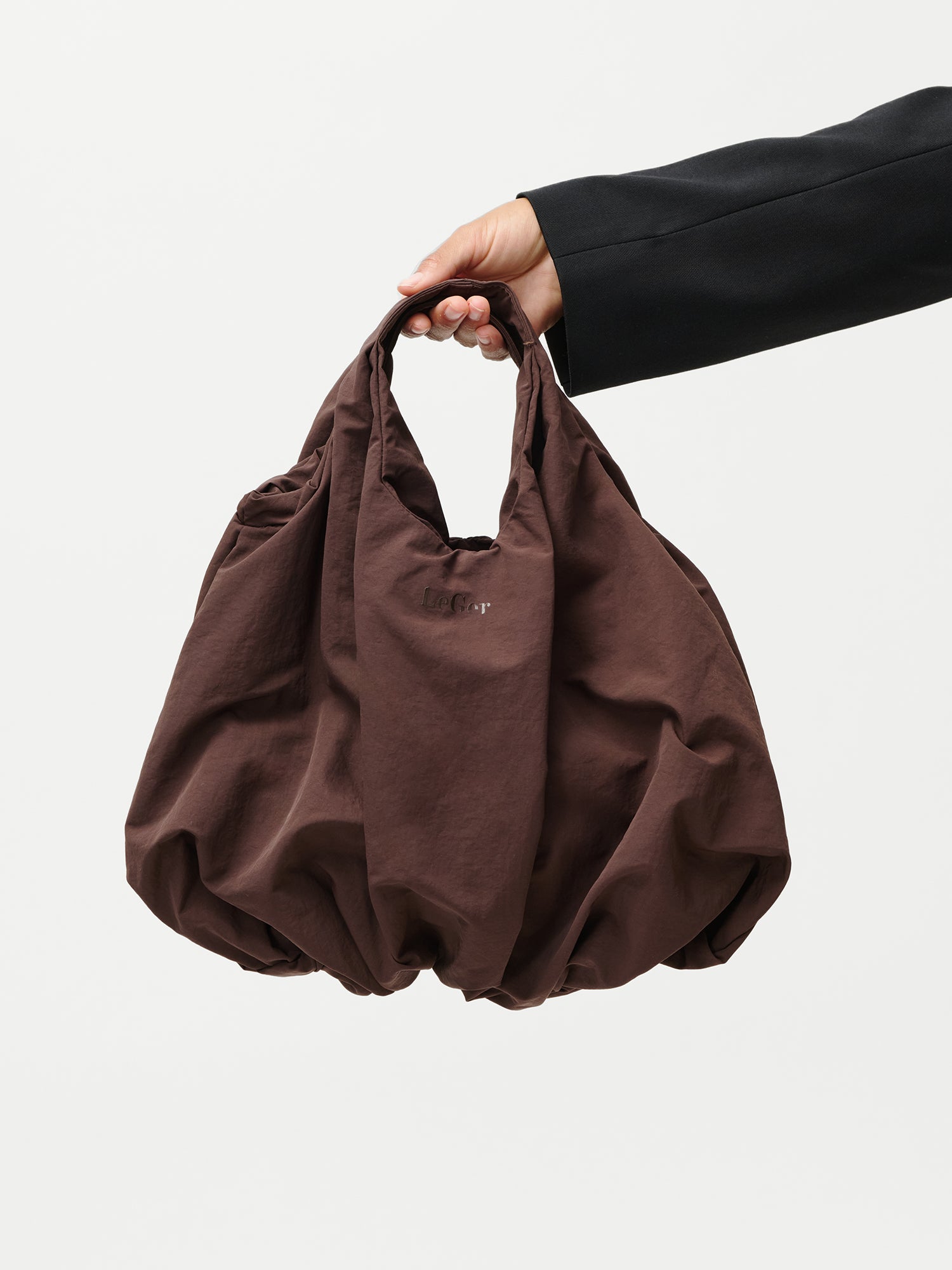 Eine Hand in einem schwarzen Ärmel hält die übergroße Lotti Bag von LeGer by Lena Gercke - braun geknittert mit gerafften Kanten und einem kleinen gestickten Logo auf der Vorderseite - vor einem schlichten weißen Hintergrund.