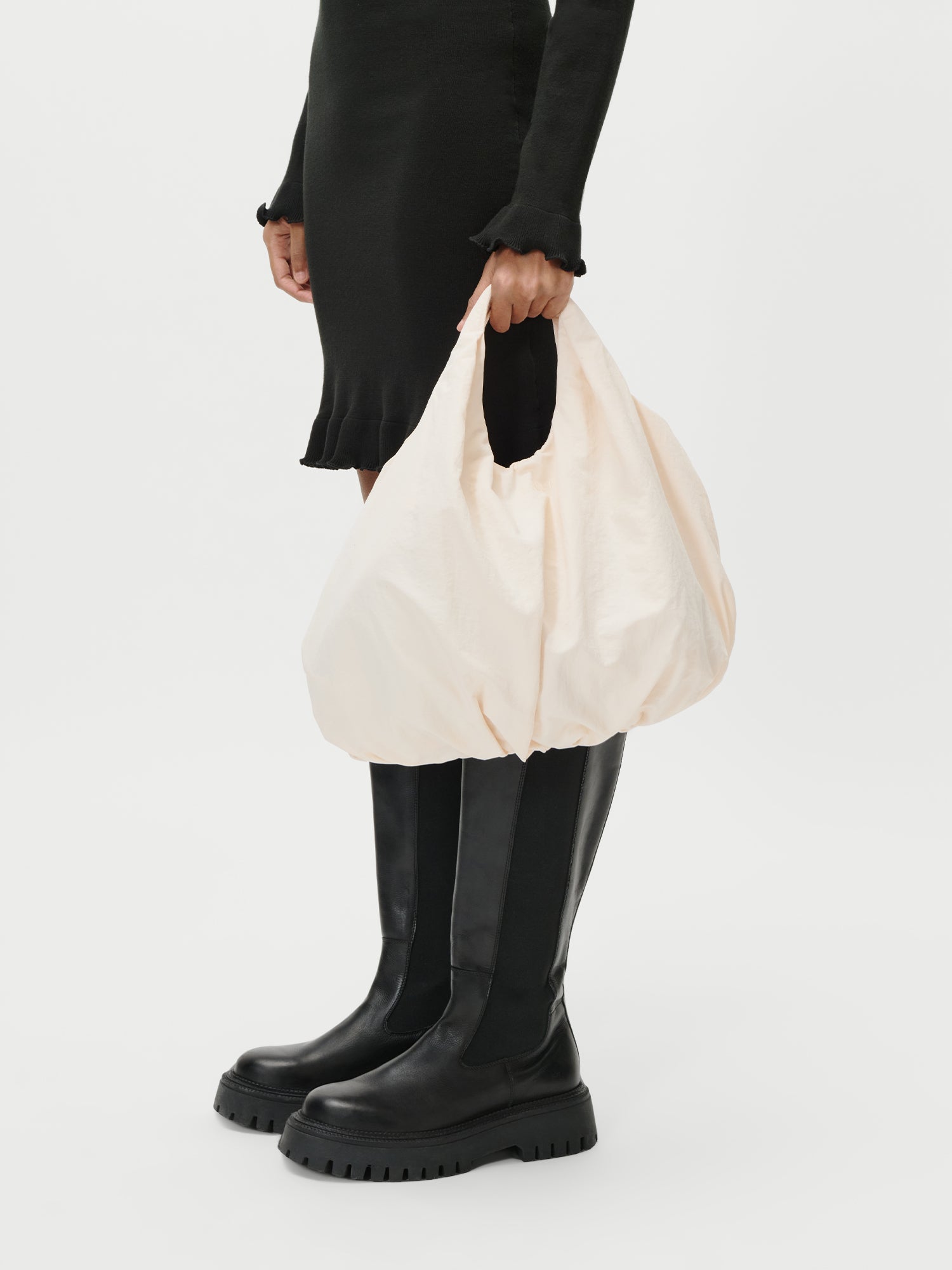 Eine Person, die ein schwarzes langärmeliges Kleid und schwarze kniehohe Stiefel trägt, hält die LeGer by Lena Gercke Lotti Bag, eine große hellbeige Tasche, an den Henkeln vor einem schlichten weißen Hintergrund.