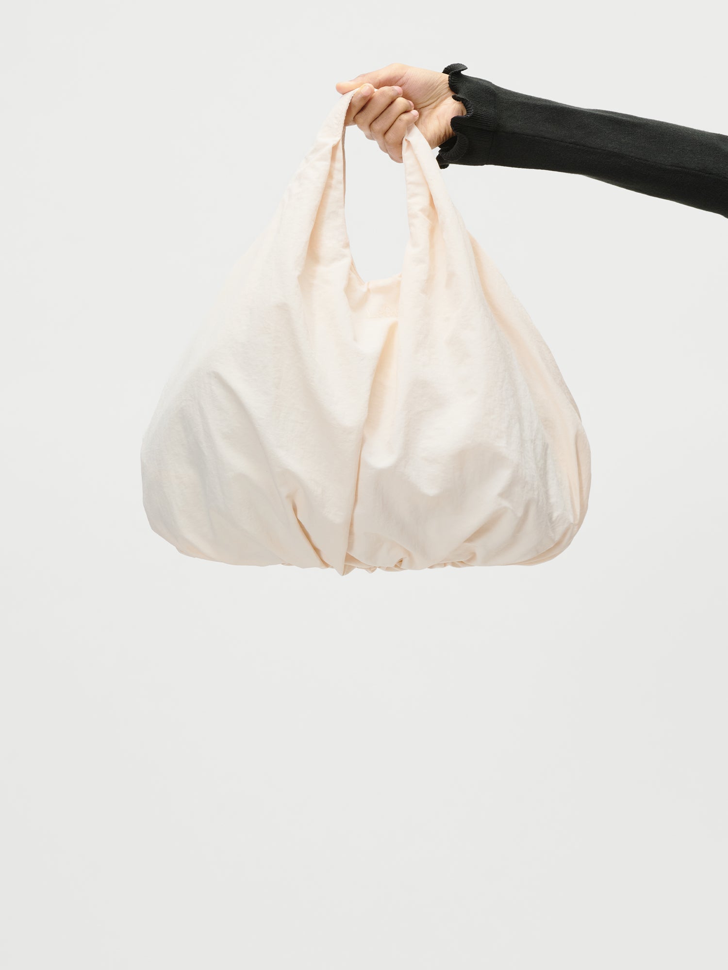 Eine Hand in einem schwarzen Ärmel hält die geräumige, weiche, cremefarbene Lotti Bag von LeGer by Lena Gercke vor einem schlichten weißen Hintergrund.