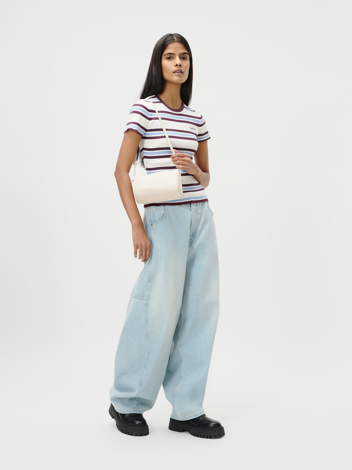 Eine Frau steht vor einem weißen Hintergrund, trägt ein gestreiftes, kurzärmeliges Oberteil, hellblaue Baggy-Jeans, schwarze, klobige Schuhe und hat die Georgia Bag von LeGer by Lena Gercke dabei.