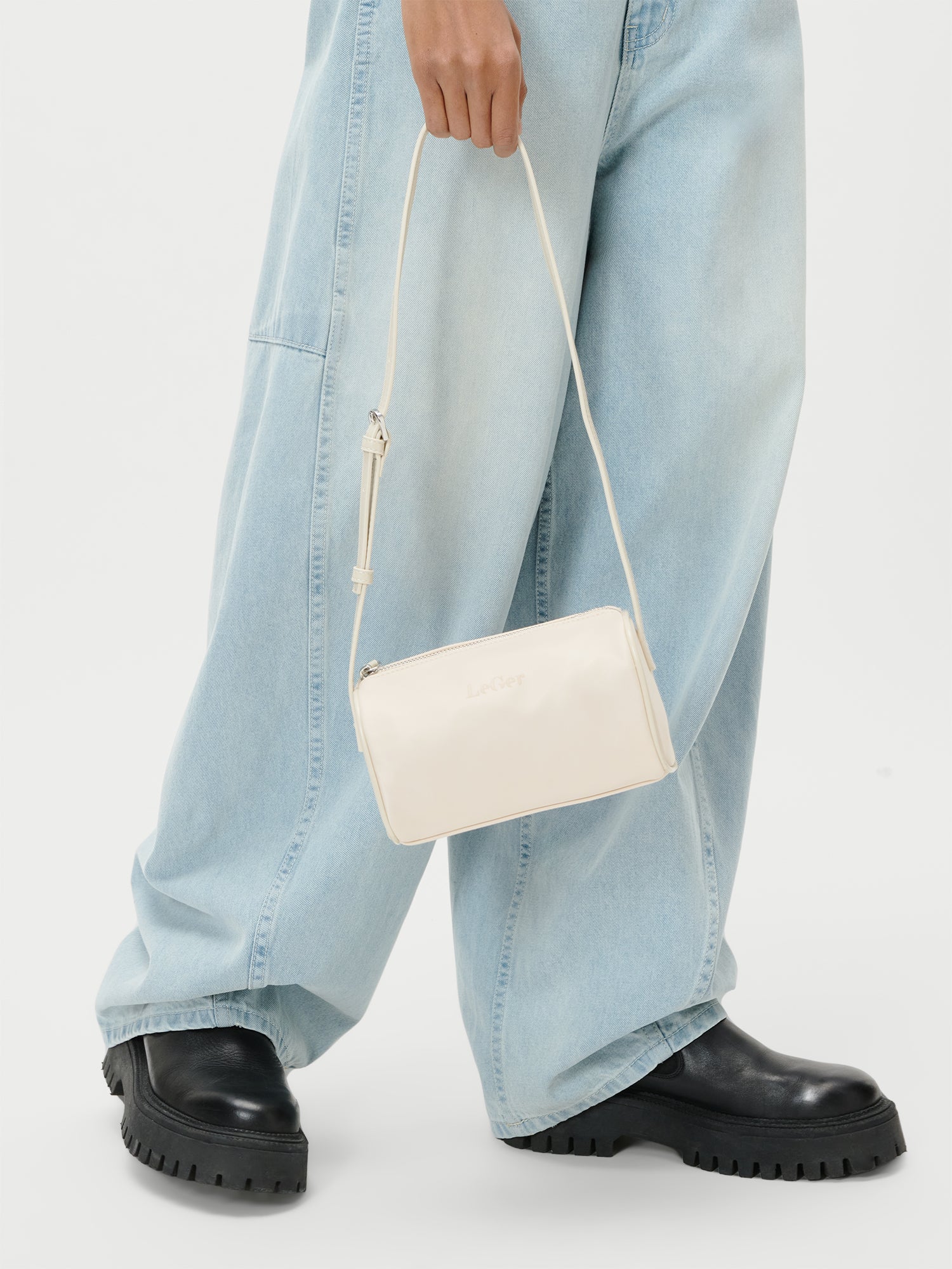 Eine Person hält die LeGer by Lena Gercke Georgia Bag - eine kleine weiße Umhängetasche - an ihrem Riemen an der Seite, während sie eine hellblaue Jeans mit weitem Bein und schwarze Schuhe mit klobigen Sohlen trägt.