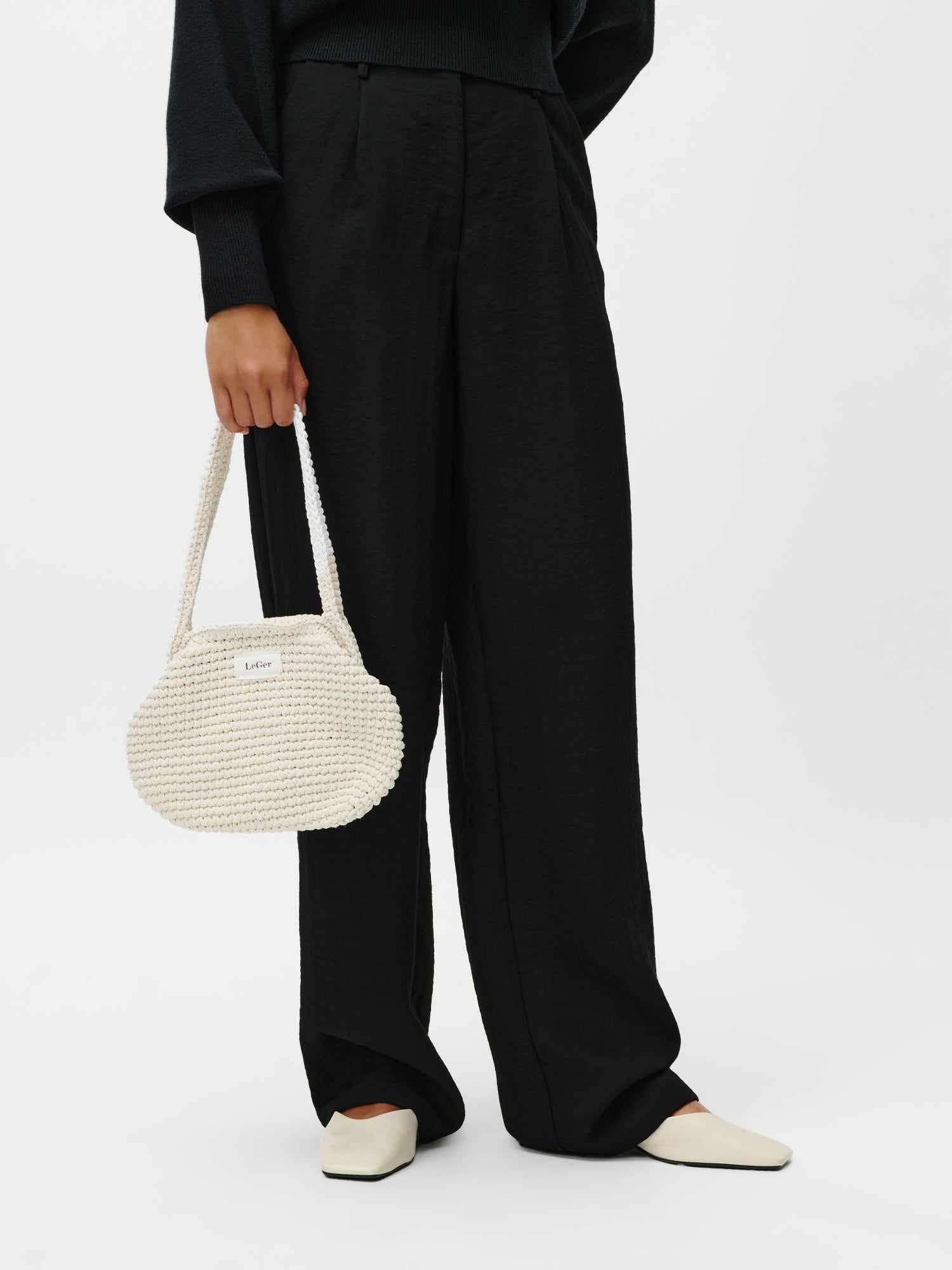 Eine Person, die eine schwarze Hose mit weitem Bein und cremefarbene Schuhe trägt, hält die LeGer by Lena Gercke Perle Bag, eine weiße strukturierte Handtasche, an ihrer Seite. Nur die untere Hälfte ist vor einem schlichten weißen Hintergrund zu sehen.