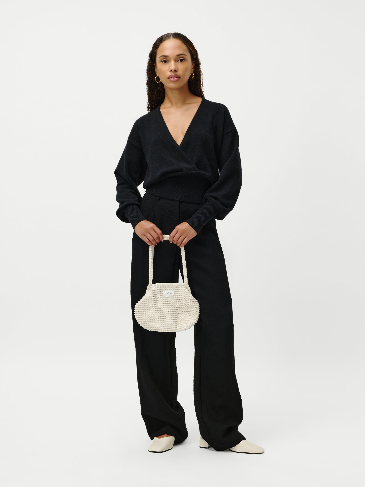 Eine Frau steht vor einem schlichten weißen Hintergrund, trägt einen schwarzen Pullover mit V-Ausschnitt, eine schwarze Hose mit weitem Bein, weiße spitze Schuhe und hält die Perle Bag von LeGer by Lena Gercke in der Hand - eine kleine runde, gewebte weiße Handtasche.