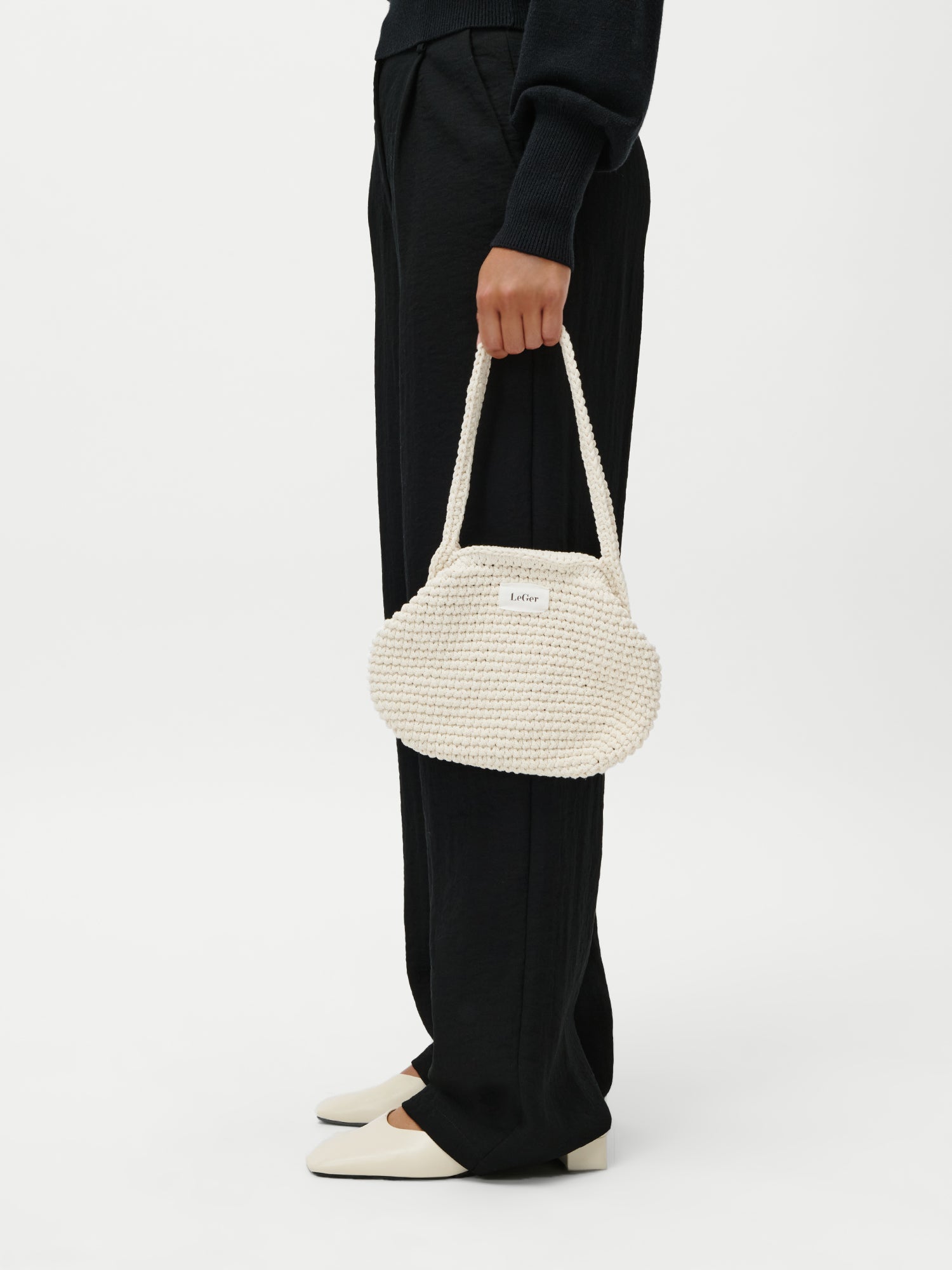 Eine Person in Schwarz hält die Perle Bag von LeGer by Lena Gercke - eine kleine, cremefarbene, geflochtene Handtasche mit einem Henkel. Sie trägt cremefarbene Schuhe mit schwarzen Sohlen und posiert vor einem schlichten weißen Hintergrund.