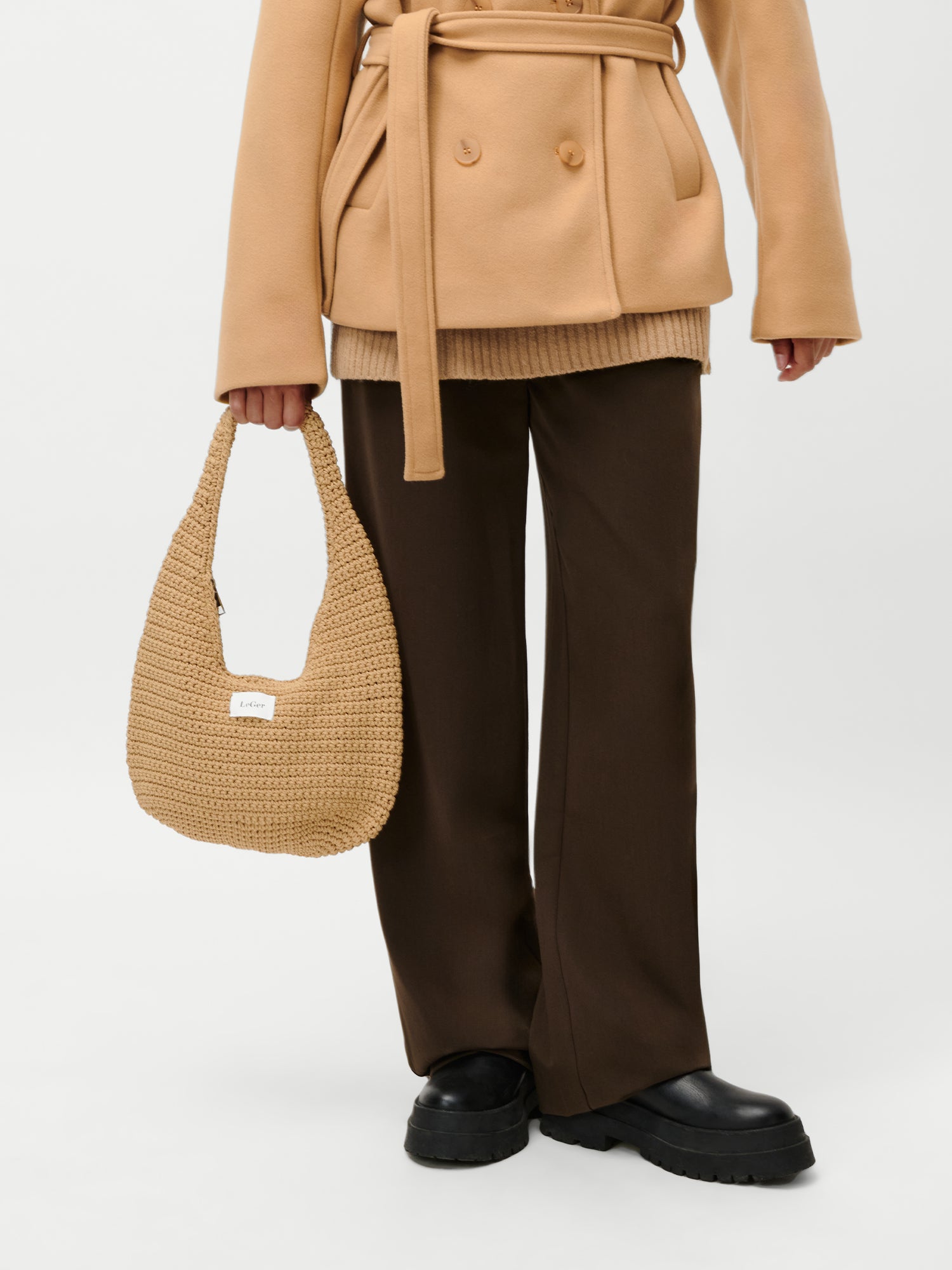 Eine Person, die einen hellbraunen Mantel mit Gürtel, eine braune Hose und schwarze Plateauschuhe trägt, hält die LeGer by Lena Gercke Theodora Bag - eine gewebte hellbraune Handtasche - in der linken Hand. Das Foto ist an den Schultern abgeschnitten, um das Outfit und die Tasche hervorzuheben.