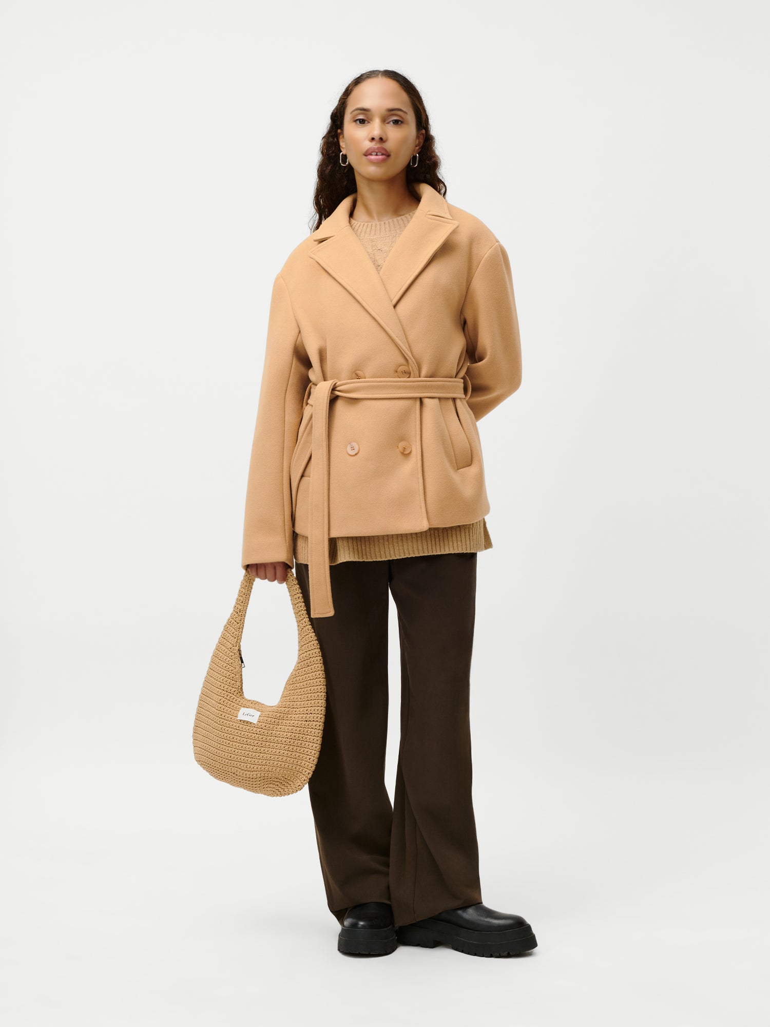 Eine Frau mit langen Haaren und einem ruhigen Gesichtsausdruck steht vor einem weißen Hintergrund und hält die LeGer by Lena Gercke Theodora Bag - eine gewebte, hellbraune Handtasche - während sie einen beigen Gürtelmantel, eine braune Hose mit weitem Bein und schwarze Schuhe trägt.