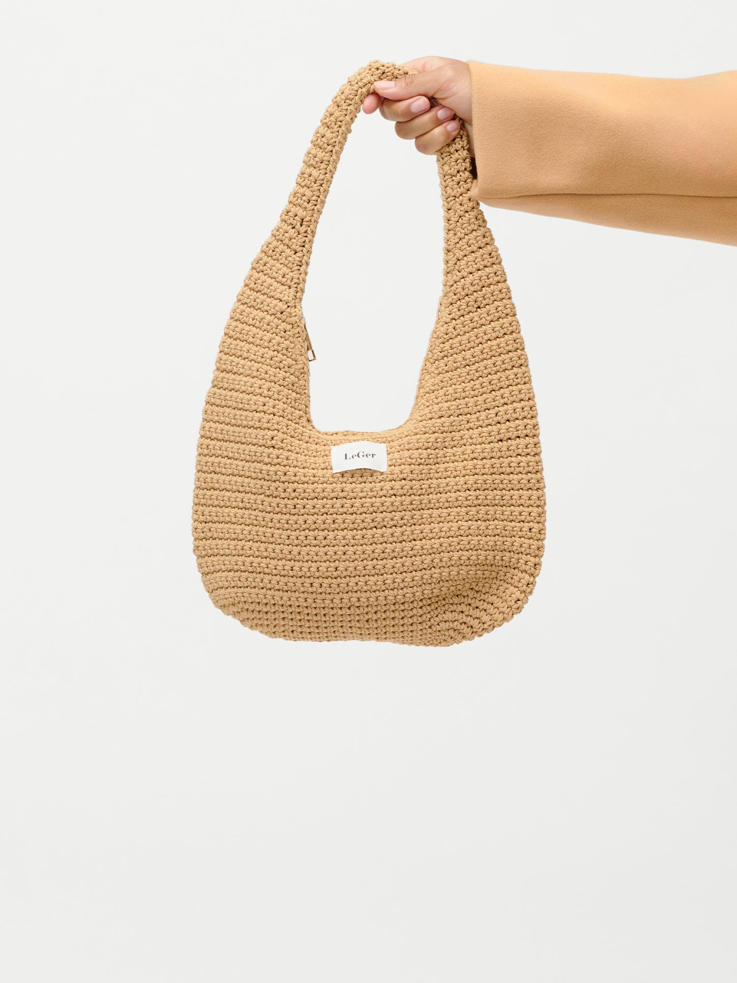 Eine Hand hält die beigefarbene, gehäkelte Theodora Bag von LeGer by Lena Gercke vor einem schlichten weißen Hintergrund. Auf der Vorderseite der Tasche ist ein Etikett mit dem Markennamen "LeGer" zu sehen.