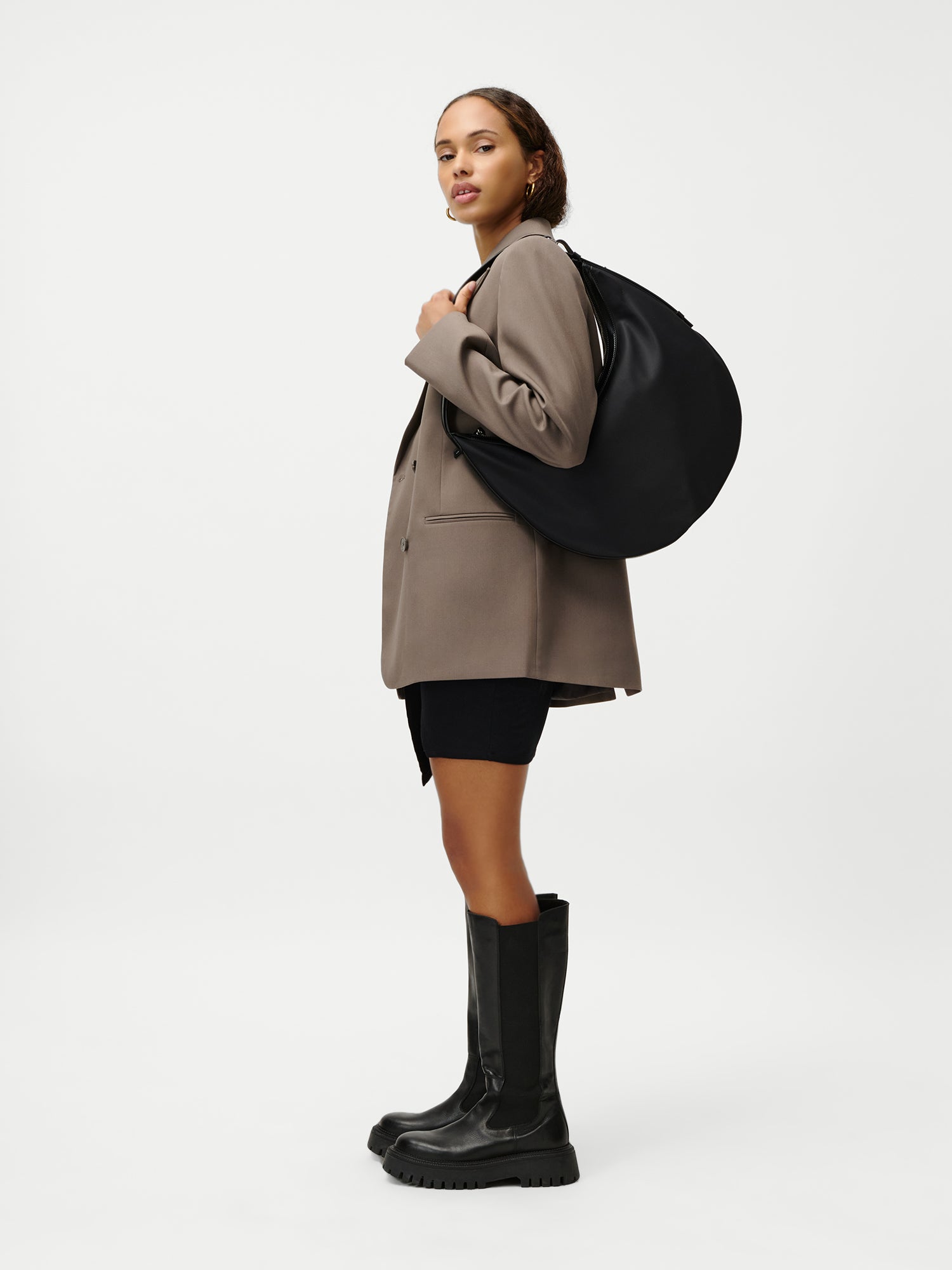 Eine Frau im Profil vor weißem Hintergrund trägt einen taupefarbenen Blazer, schwarze Shorts, kniehohe schwarze Stiefel und trägt die Sarina Bag von LeGer by Lena Gercke - eine große schwarze Tasche, die sie sich über die Schulter hängt.
