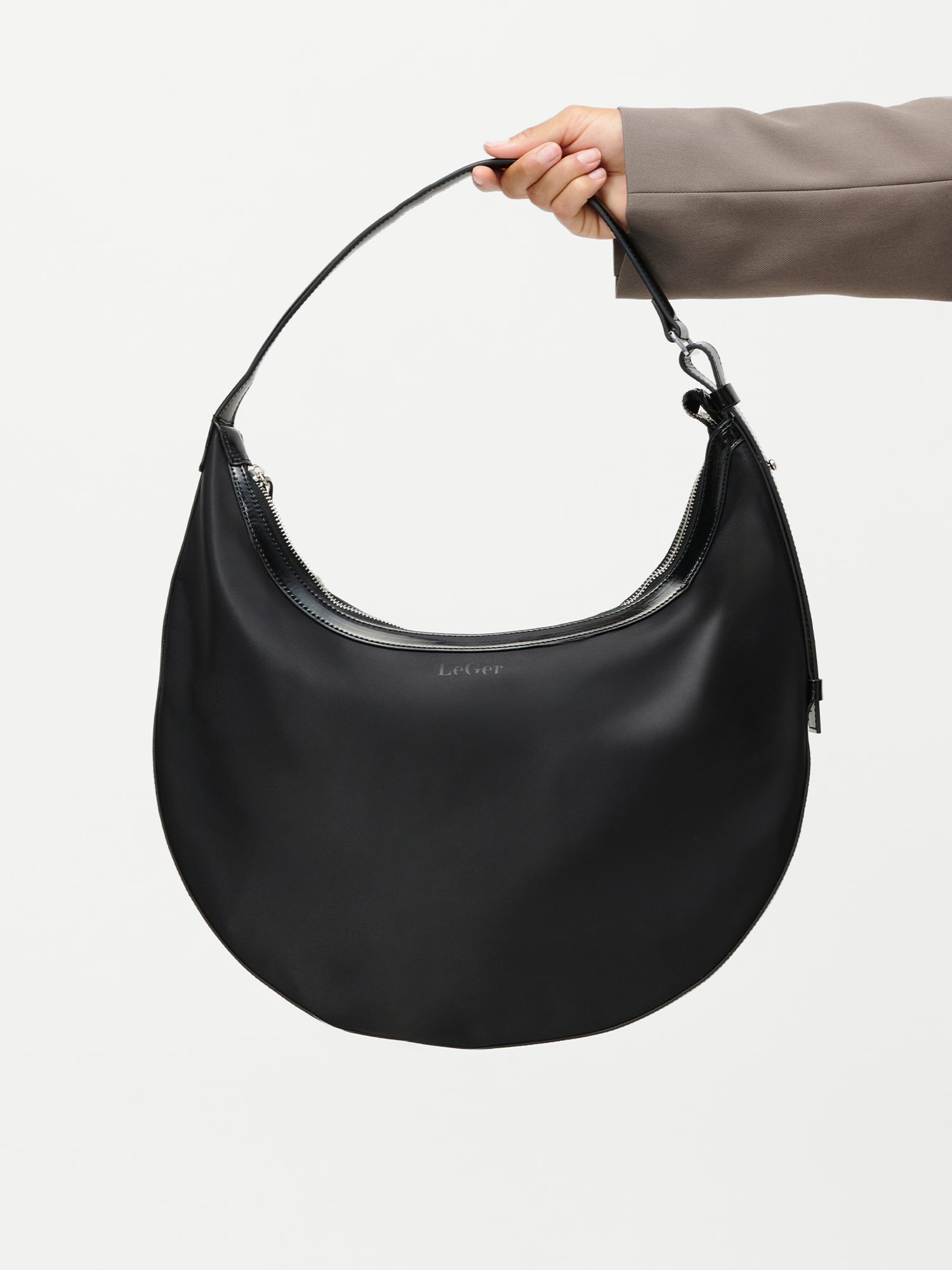 Eine Person in einem taupefarbenen Ärmel hält die LeGer by Lena Gercke Sarina Bag, eine große, sichelförmige schwarze Handtasche mit einem einzigen Riemen und Reißverschluss, vor einem schlichten weißen Hintergrund.