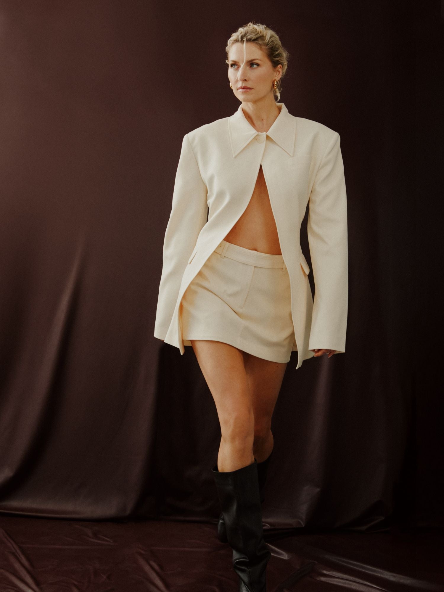 Eine Frau posiert in einem cremefarbenen Cim Blazer von LeGer by Lena Gercke, gepaart mit einem passenden Minirock und schwarzen kniehohen Stiefeln vor einem dunkelbraunen Hintergrund. Der offene Blazer im Brustbereich setzt ein mutiges, stilvolles Statement.