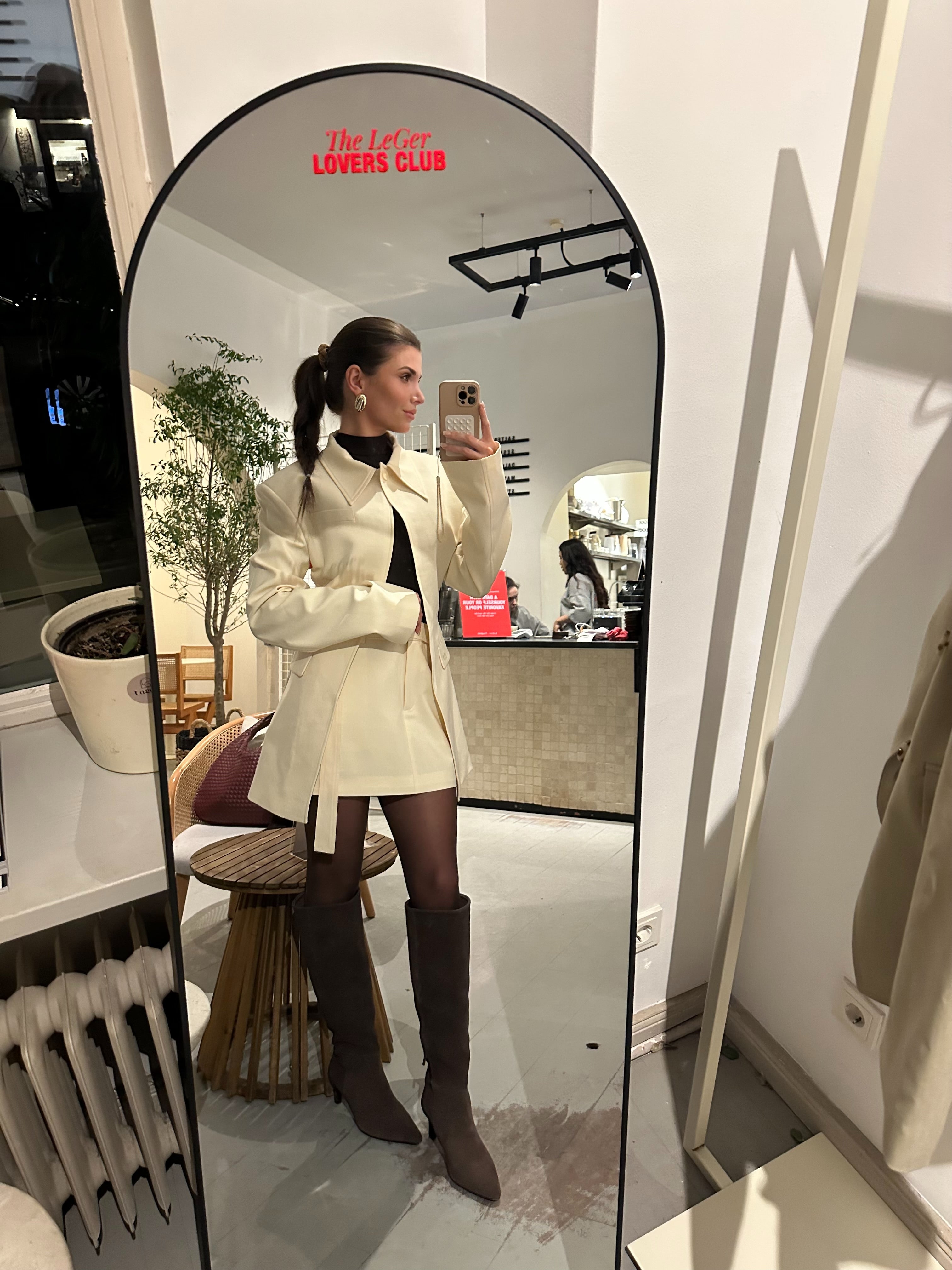 Eine Frau, die den Cim Blazer von LeGer by Lena Gercke trägt, macht ein Spiegel-Selfie in einem modernen, mit Pflanzen gefüllten Raum und kombiniert die cremefarbene Jacke mit braunen, kniehohen Stiefeln, einer schwarzen Strumpfhose und einem weißen Hemd.