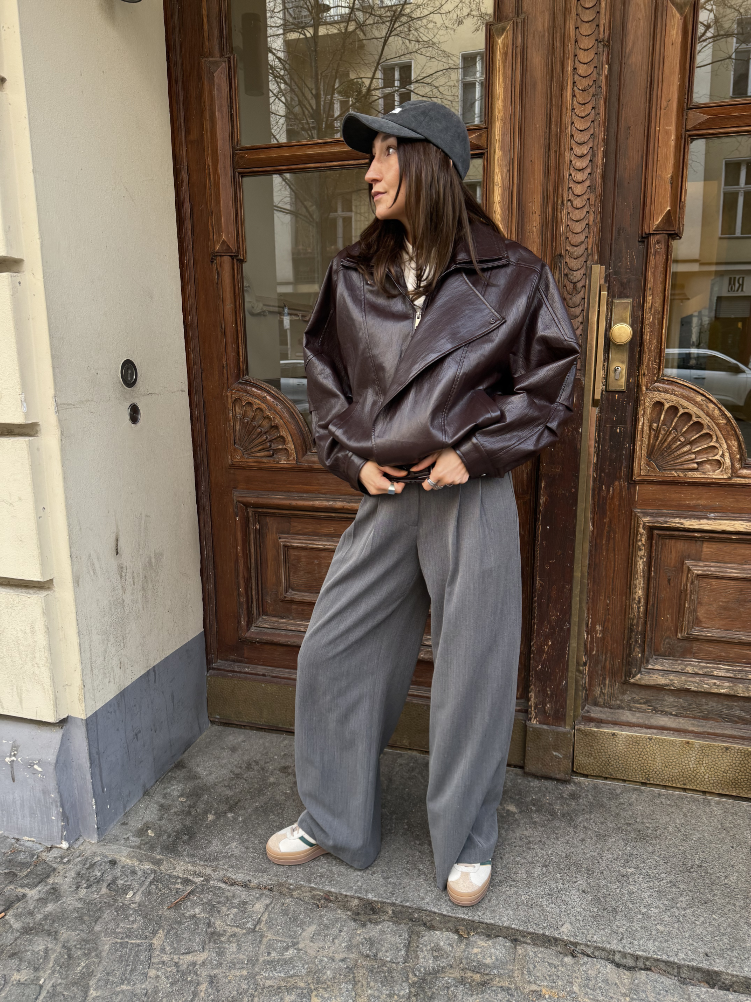 Eine Frau steht vor einer Holztür und trägt die Jacke Jessa von LeGer by Lena Gercke, kombiniert mit einer weiten grauen Hose, beigen Turnschuhen und einer grauen Mütze. Die Hände stecken in den Taschen, während sie nach links schaut.