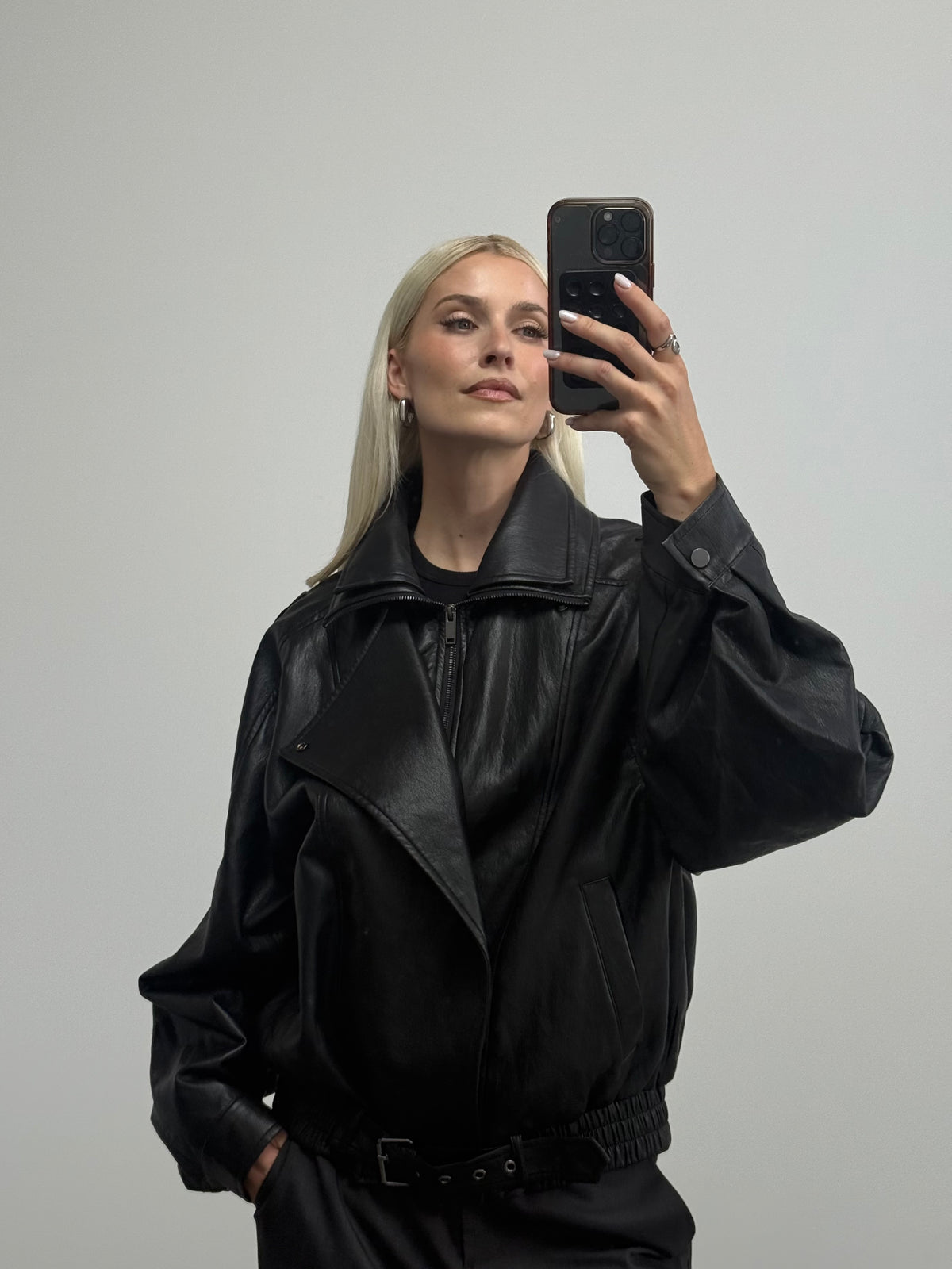 Eine Person mit langen blonden Haaren macht ein Spiegel-Selfie und präsentiert dabei einen Look im Bikerstil: Sie trägt die übergroße „Jessa“-Jacke von LeGer by Lena Gercke und eine schwarze Hose vor einem schlichten hellgrauen Hintergrund.