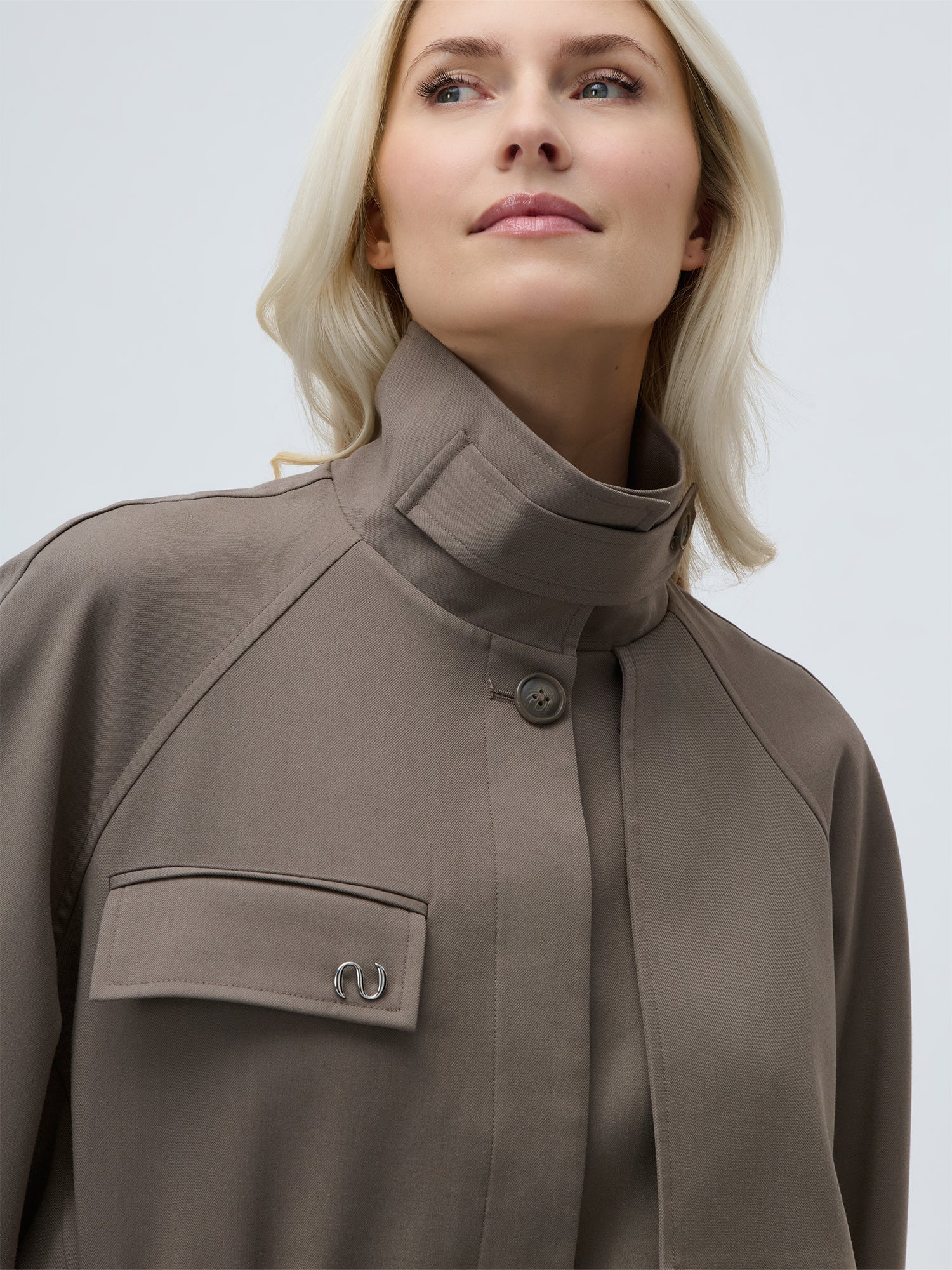 Eine Frau mit langen blonden Haaren trägt die Fee Jacket by LeGer by Lena Gercke in taupe - eine locker sitzende Damenjacke mit hohem Kragen, geknöpfter Vorderseite und Pattentasche, die vor einem schlichten hellen Hintergrund selbstbewusst nach oben schaut.