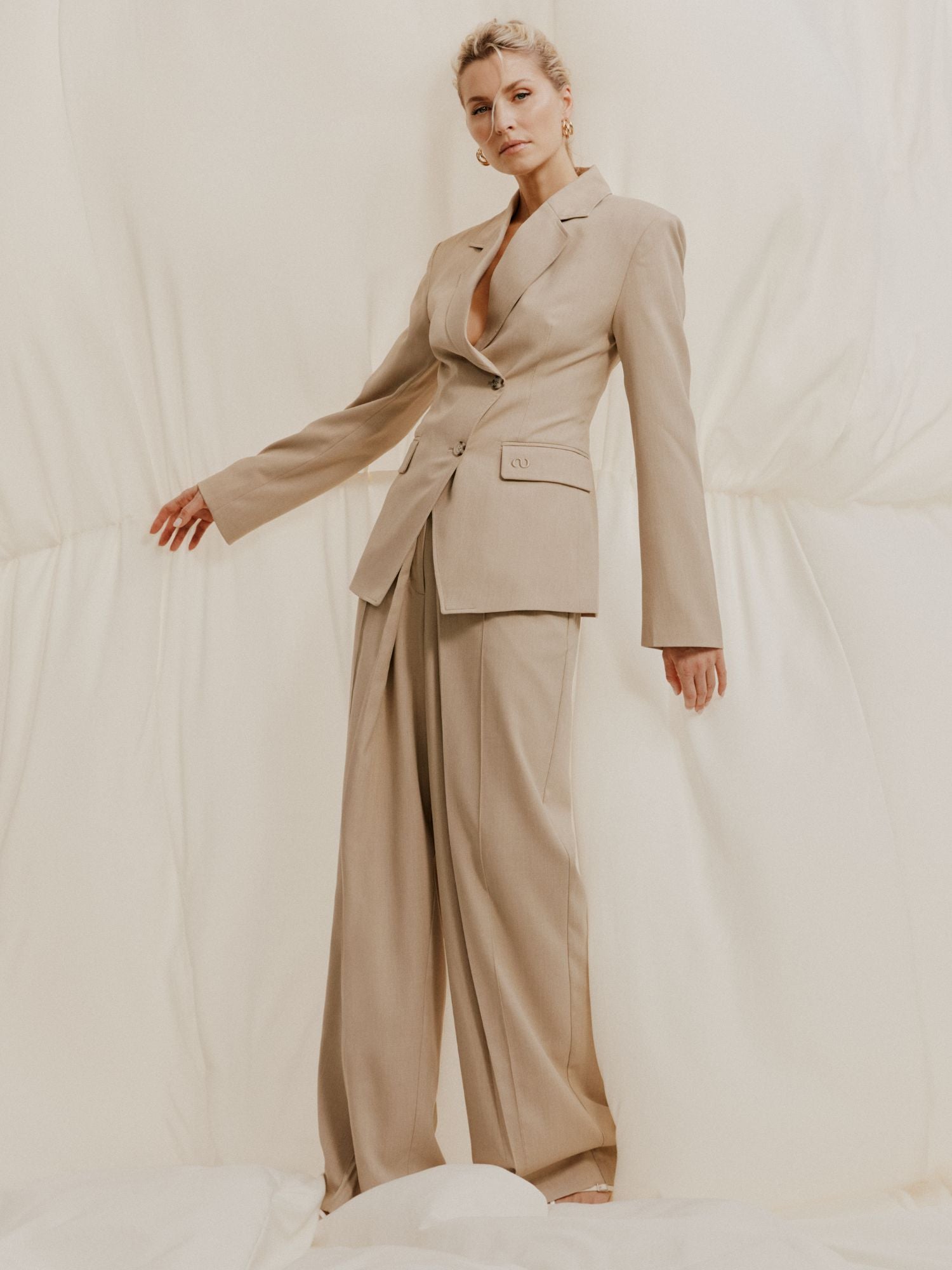 Eine Frau posiert selbstbewusst vor einem cremefarbenen Hintergrund. Sie trägt die Raquel Pants von LeGer by Lena Gercke - eine locker sitzende Hose mit weitem Bein und hoher Taille - und dazu einen maßgeschneiderten Blazer. Ihr Haar ist zurückgesteckt und ein Arm ist leicht ausgestreckt.