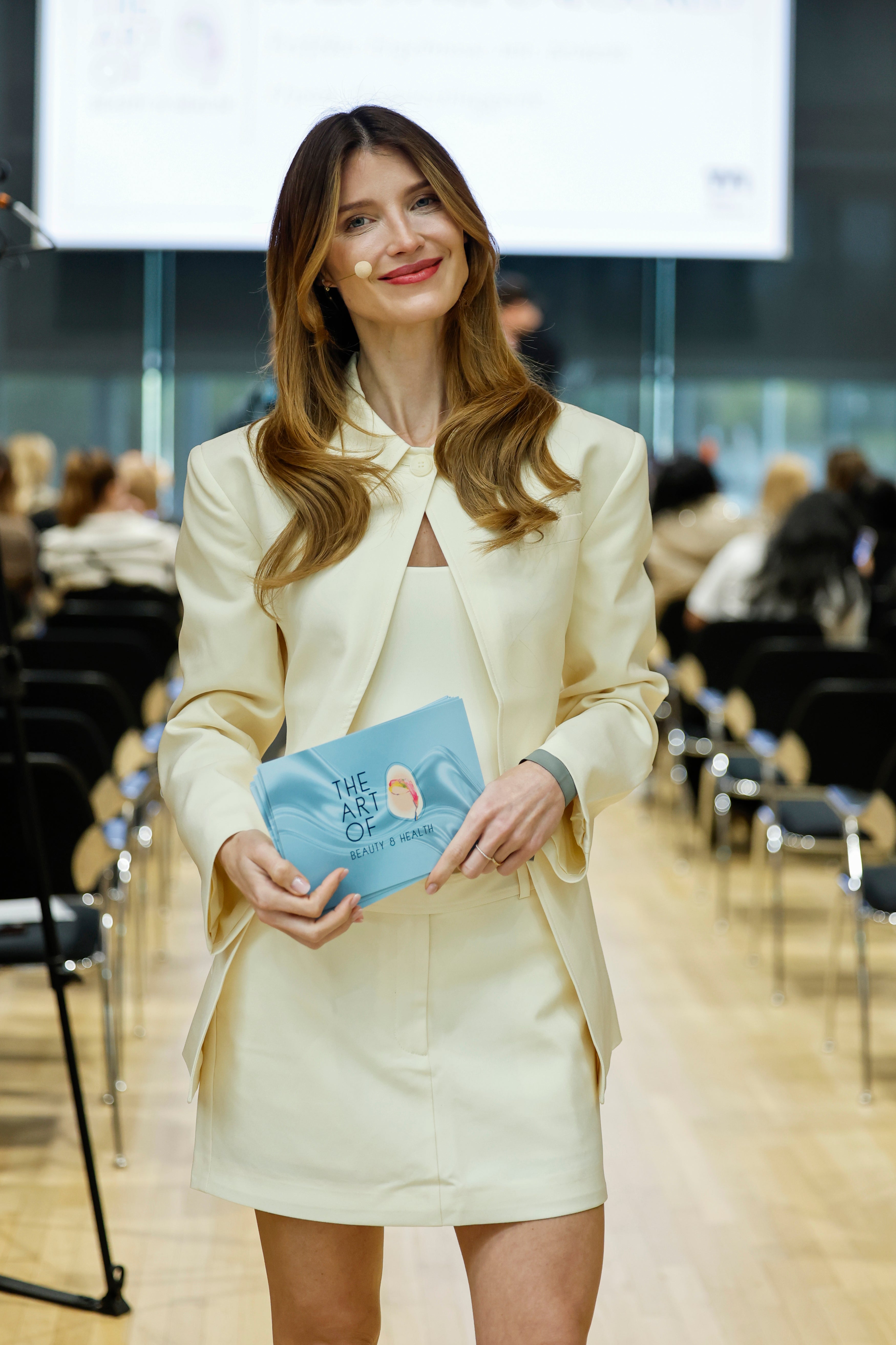Eine Frau mit langem, gewelltem Haar steht in einem Haus, lächelt und hält ein Buch in der Hand. Sie trägt einen cremefarbenen mittellangen Cim-Rock von LeGer by Lena Gercke und einen Blazer. Eine Reihe von Menschen im Hintergrund deutet auf eine formelle Veranstaltung oder Präsentation hin.