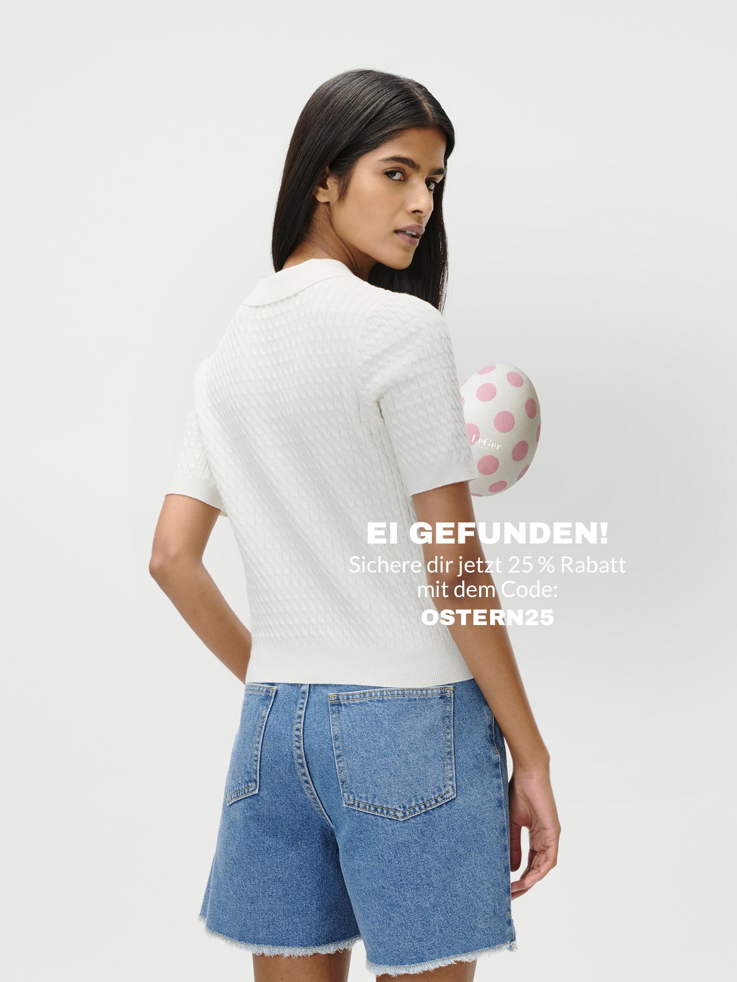 Eine Frau mit langen dunklen Haaren trägt das Manolja T-Shirt von LeGer by Lena Gercke in Größe 36 und Denim Shorts, hält ein pinkes Polka-Dot-Ei hinter dem Rücken. Spare 25 % mit Code OSTERN25 - jetzt auf Deutsch!.