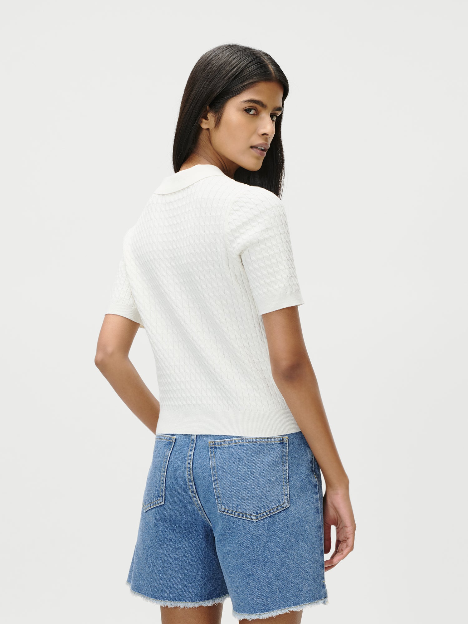 Eine Frau mit langen dunklen Haaren, die wie Lena Gercke gestylt ist, trägt das Manolja T-Shirt von LeGer by Lena Gercke in Weiß, gepaart mit hochgeschlossenen blauen Jeansshorts, vor einem einfarbig weißen Hintergrund und schaut über ihre Schulter.