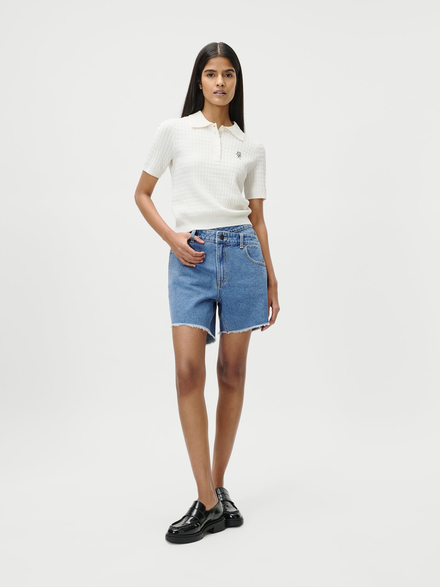 Eine Frau in Größe 36 trägt das weiße Manolja T-Shirt von LeGer by Lena Gercke, blaue kurze Denim-Shorts und schwarze Schuhe. Sie steht vor weißem Hintergrund mit einer Hand in der Tasche und neutralem Gesichtsausdruck.