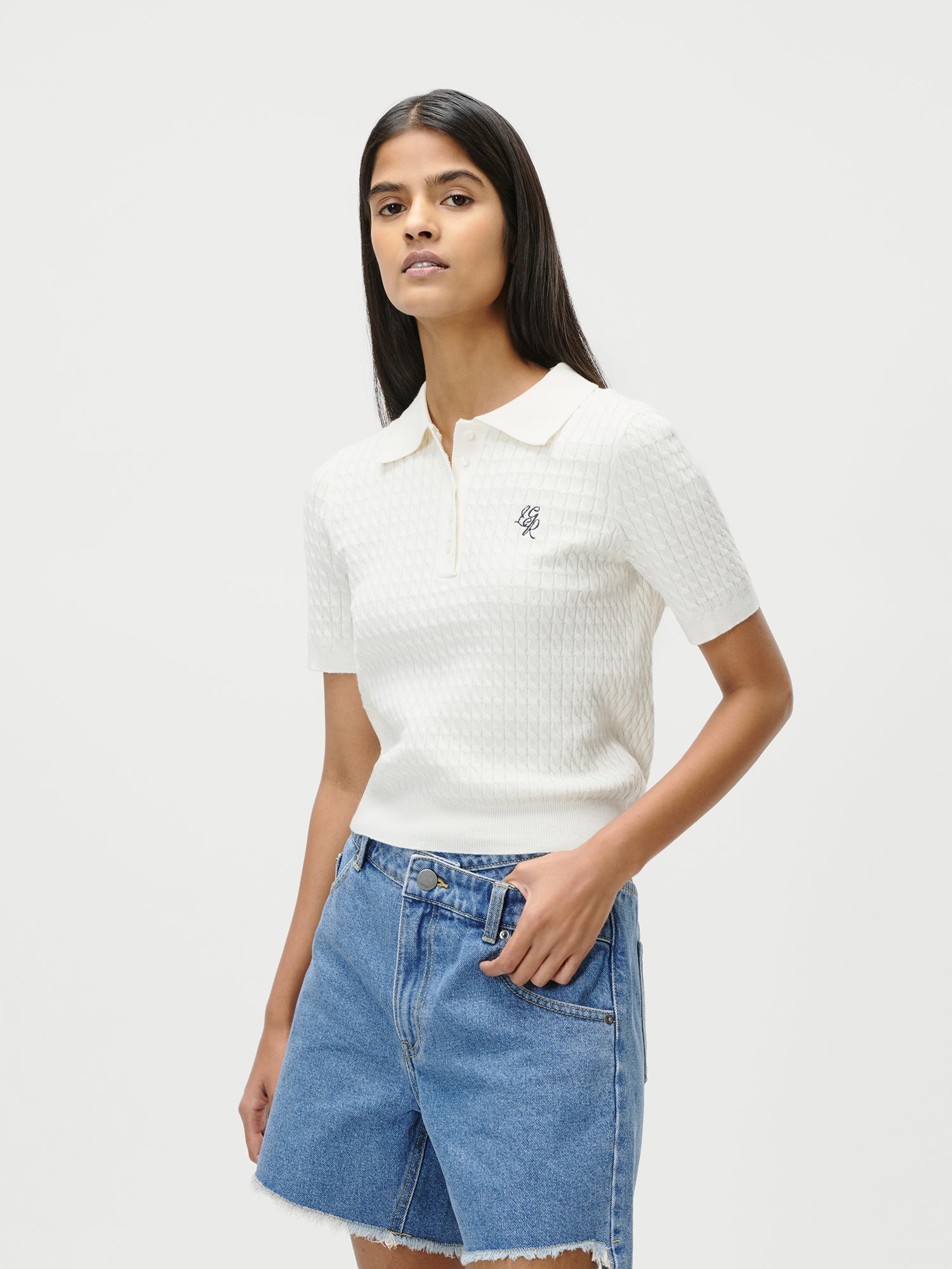 Eine junge Frau mit langen dunklen Haaren trägt das Manolja T-Shirt von LeGer by Lena Gercke in Weiß, Größe 36, und hochgeschnittene blaue Jeansshorts. Sie steht vor einem schlichten weißen Hintergrund und blickt selbstbewusst in die Kamera.