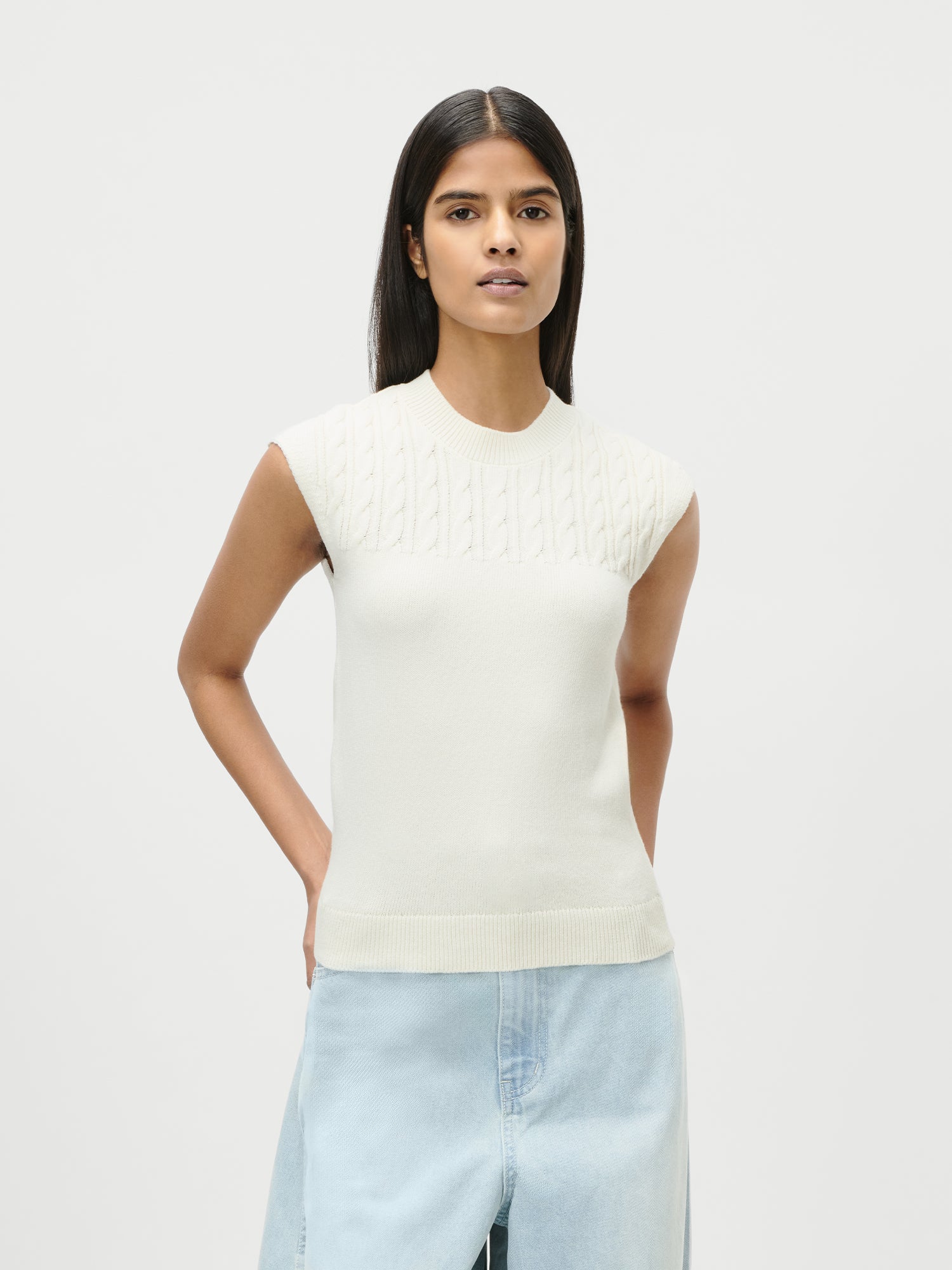 Eine Frau mit langen dunklen Haaren trägt das cremefarbene Nastasja Top von LeGer by Lena Gercke, einen kurzärmeligen Strickpullover mit Zopfmuster, kombiniert mit einem hellblauen Jeansrock. Abgebildet in Größe 36 vor einem schlichten weißen Hintergrund.