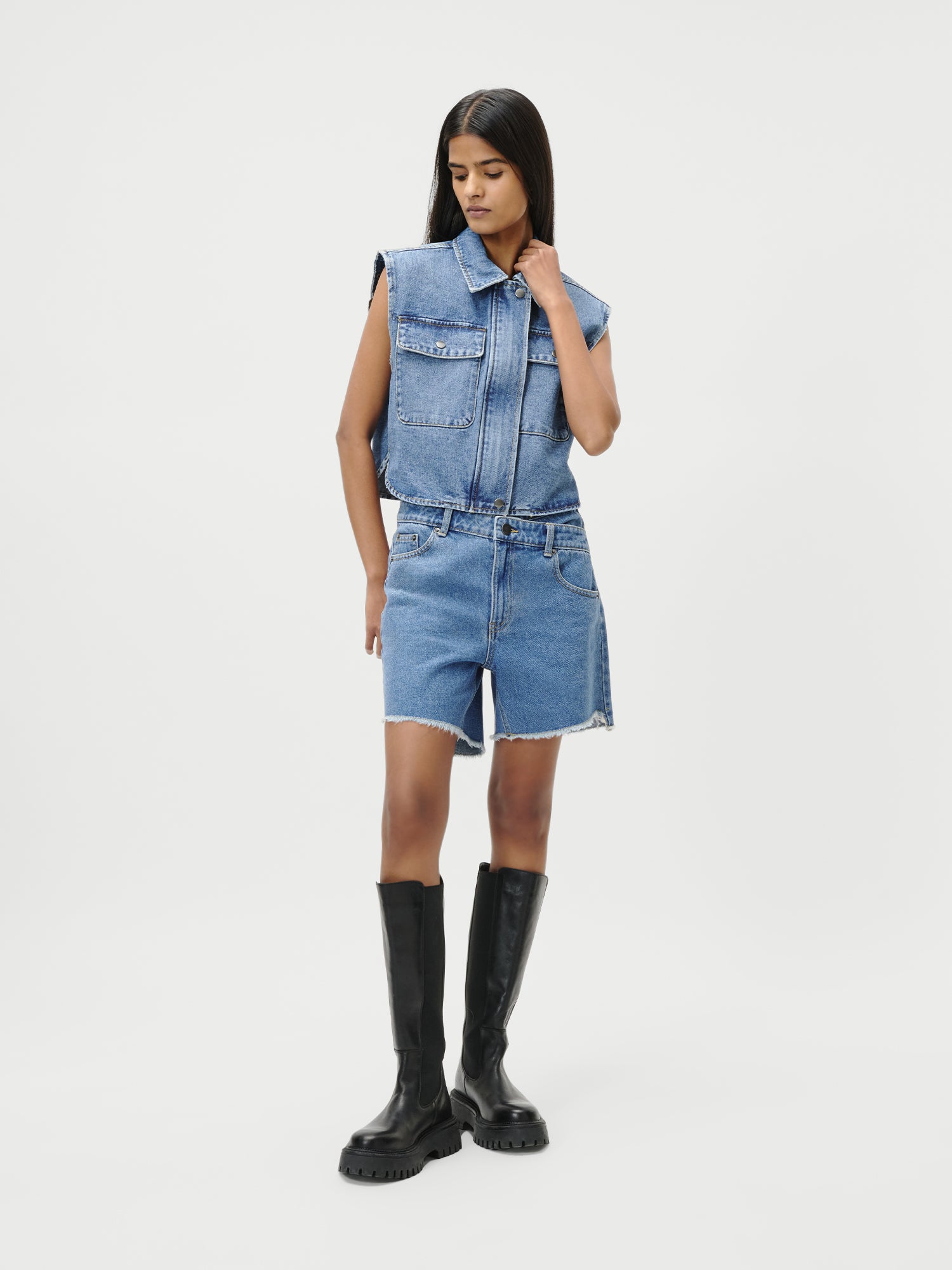 Eine Frau (Größe 36) posiert vor einem weißen Hintergrund und trägt die LeGer by Lena Gercke Shirin Vest mit Jeansshorts und kniehohen schwarzen Stiefeln, eine Hand im Haar, den Kopf leicht nach unten geneigt. Passform: Normal.