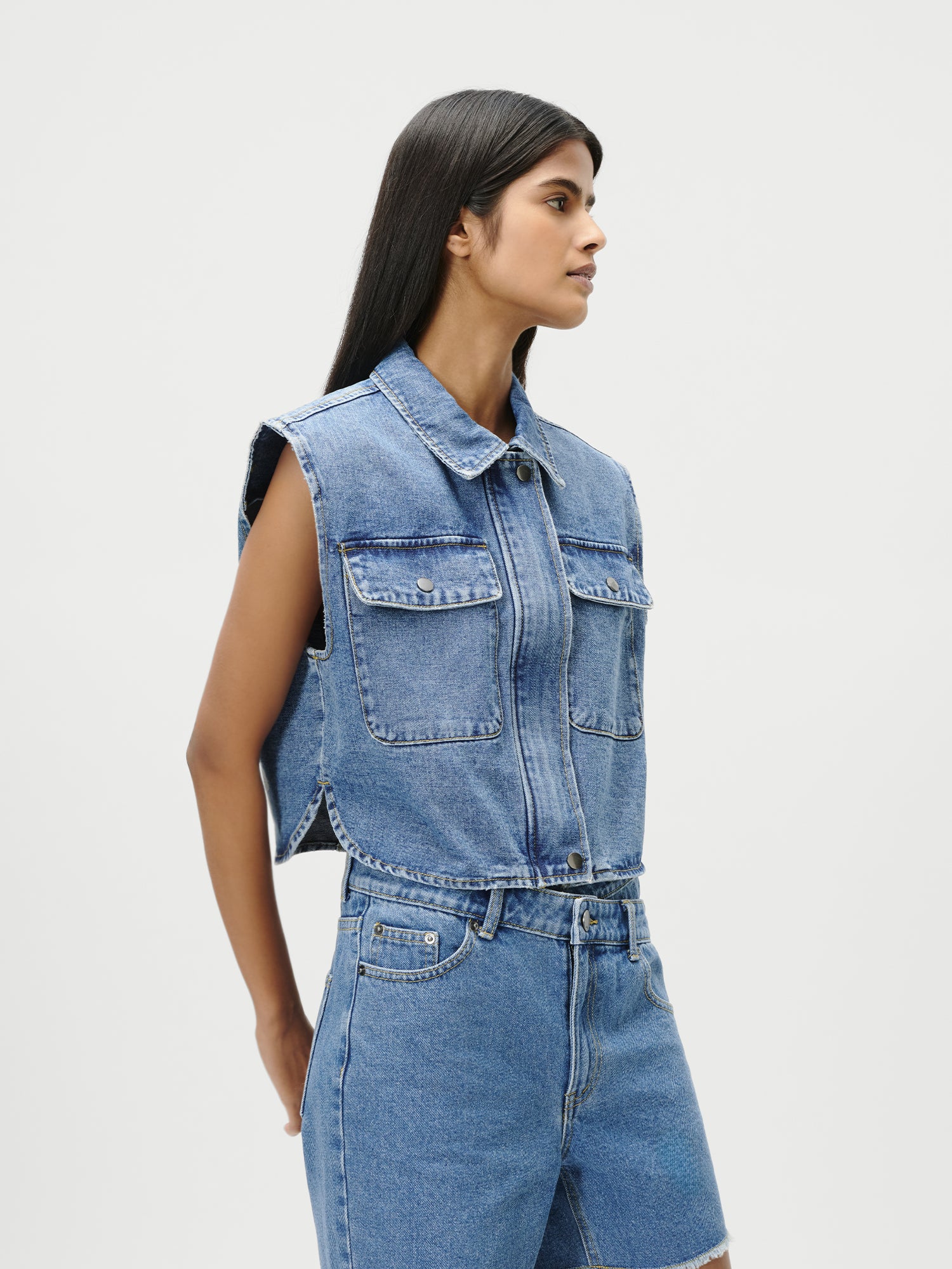 Eine Frau mit langen dunklen Haaren trägt die LeGer by Lena Gercke Shirin Vest, ein ärmelloses Jeansoberteil mit großen Taschen auf der Vorderseite, gepaart mit passenden High-Waisted-Shorts in Größe 36, vor einem schlichten weißen Hintergrund.