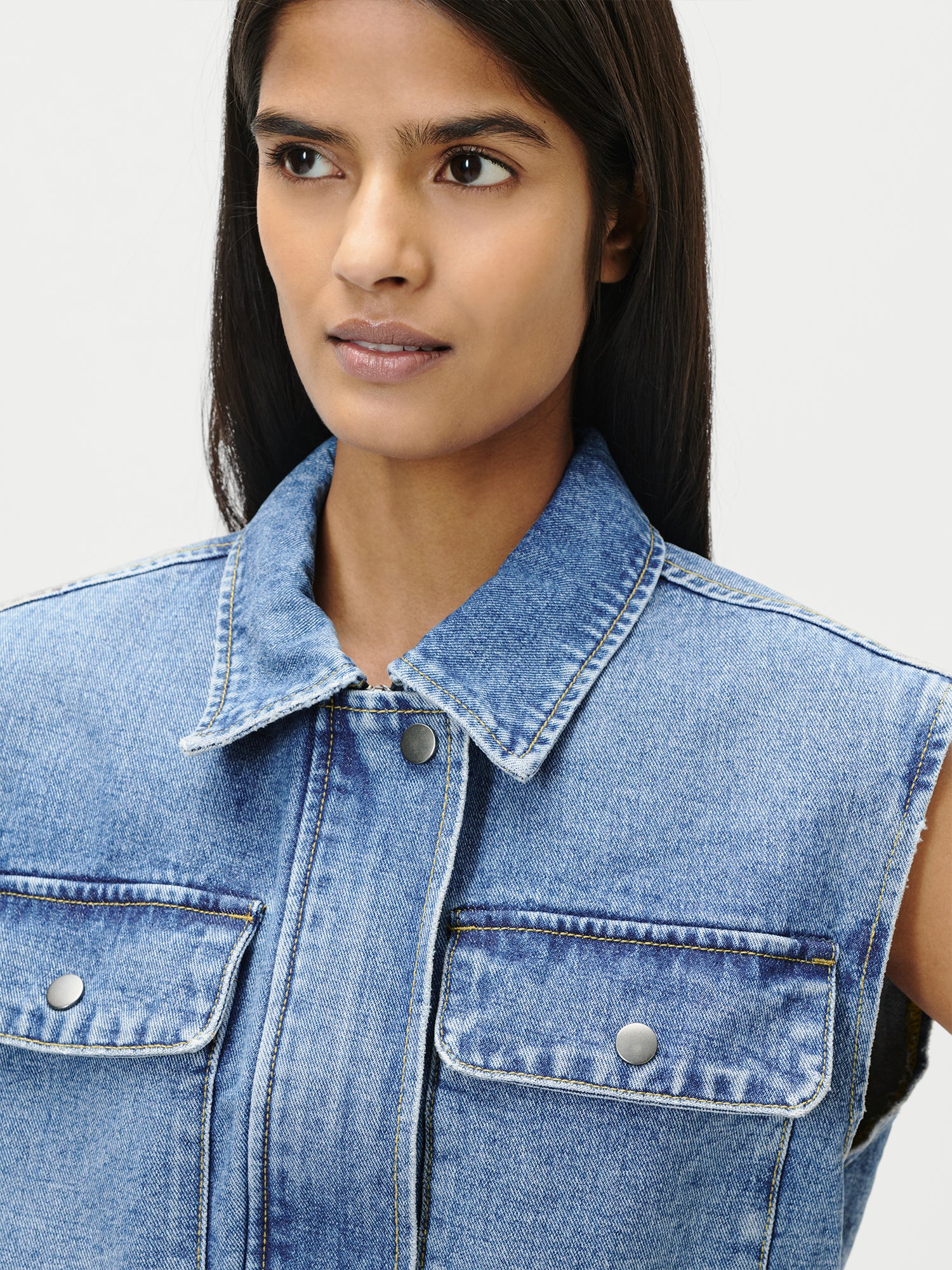 Eine Person mit glattem dunklem Haar modelliert die Shirin Vest von LeGer by Lena Gercke in Größe 36 - eine ärmellose blaue Jeansjacke mit Pattentaschen und Metallknöpfen - und schaut dabei leicht zur Seite vor einem schlichten hellen Hintergrund.