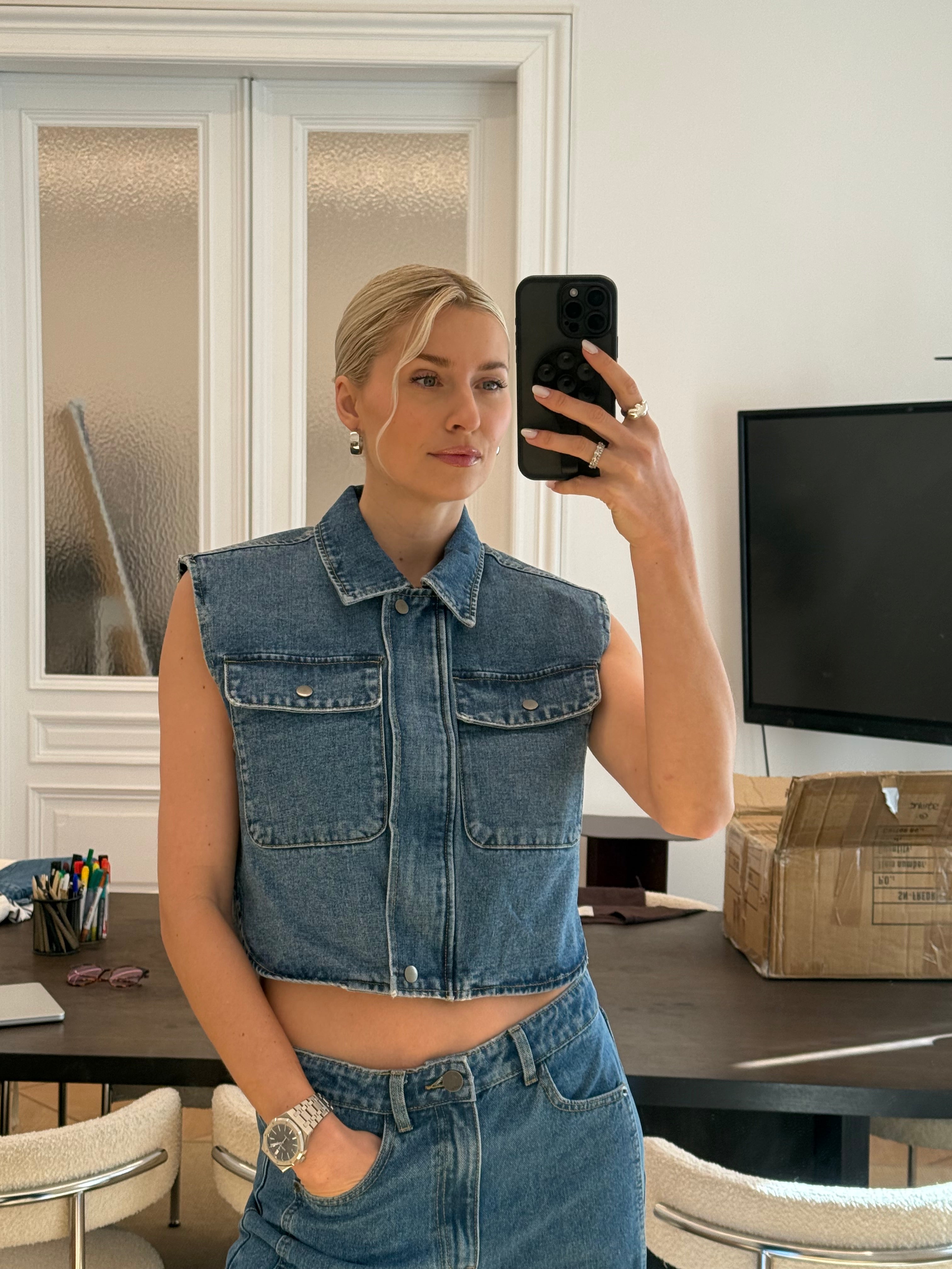 Eine Person mit blondem Haar in einem tiefen Dutt macht ein Selfie im Spiegel und trägt die Shirin Vest von LeGer by Lena Gercke und eine passende Jeanshose in Größe 36. Auf einem Tisch hinter ihnen sind Büromaterial und ein Karton zu sehen. Passform: normal.