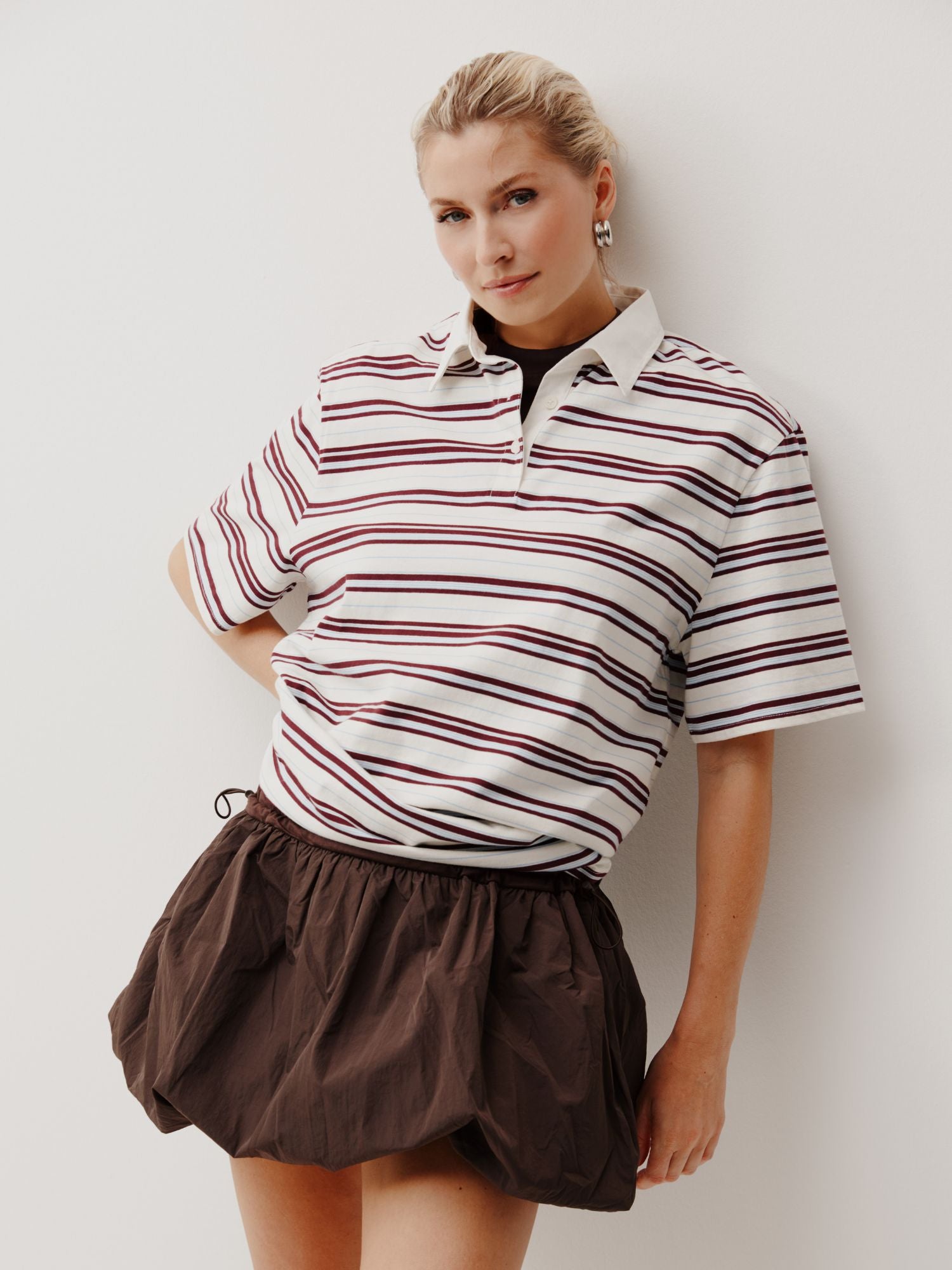 Eine Frau mit blondem Haar posiert vor einer weißen Wand und trägt das Cora T-Shirt von LeGer by Lena Gercke, ein weißes Kragenhemd mit kastanienbraunen Streifen, gepaart mit einem kurzen braunen Puff-Rock. Sie schaut mit entspanntem Blick in die Kamera.