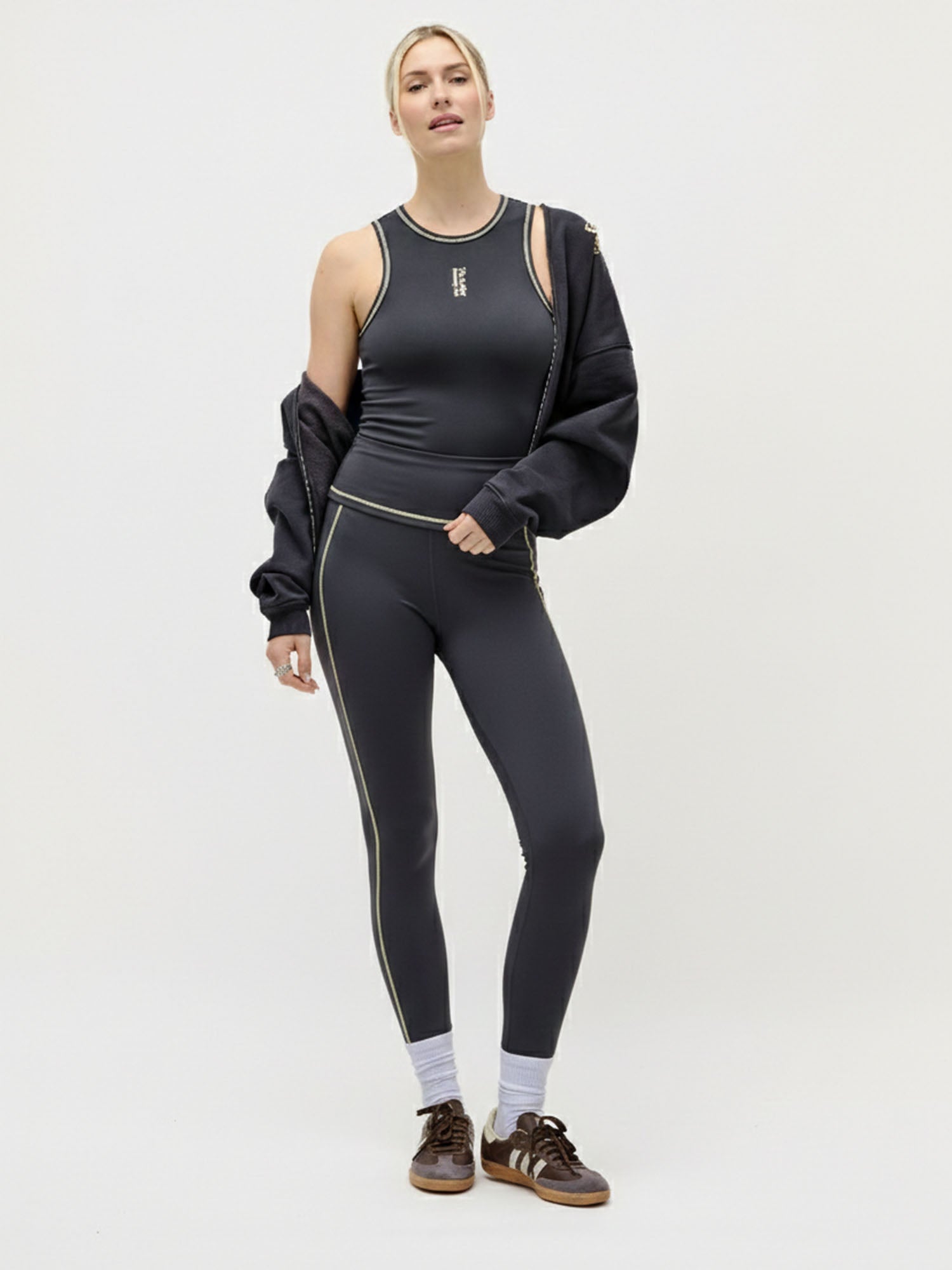 Eine Frau steht vor einem unifarbenen Hintergrund und trägt die Robinia Strumpfhose von LeGer by Lena Gercke, ein schwarzes Sporttop mit weißem Saum, eine schwarze Jacke, die über ihre Arme gezogen ist, weiße Socken und braune Turnschuhe.