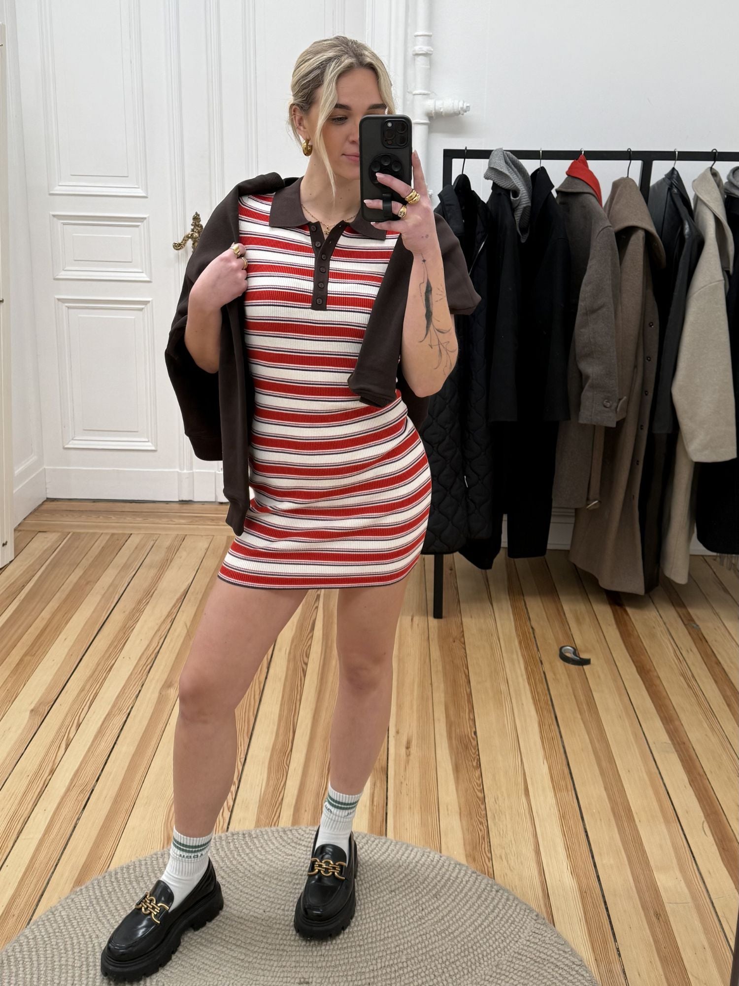 Eine Frau macht ein Spiegel-Selfie in einem Raum mit Holzboden. Sie trägt das Elenia-Kleid von LeGer by Lena Gercke mit rot-weißem Streifenmuster, dazu schwarze Slipper, weiße Socken und einen braunen Pullover über den Schultern. Hinter ihr hängen Mäntel an einem Regal.