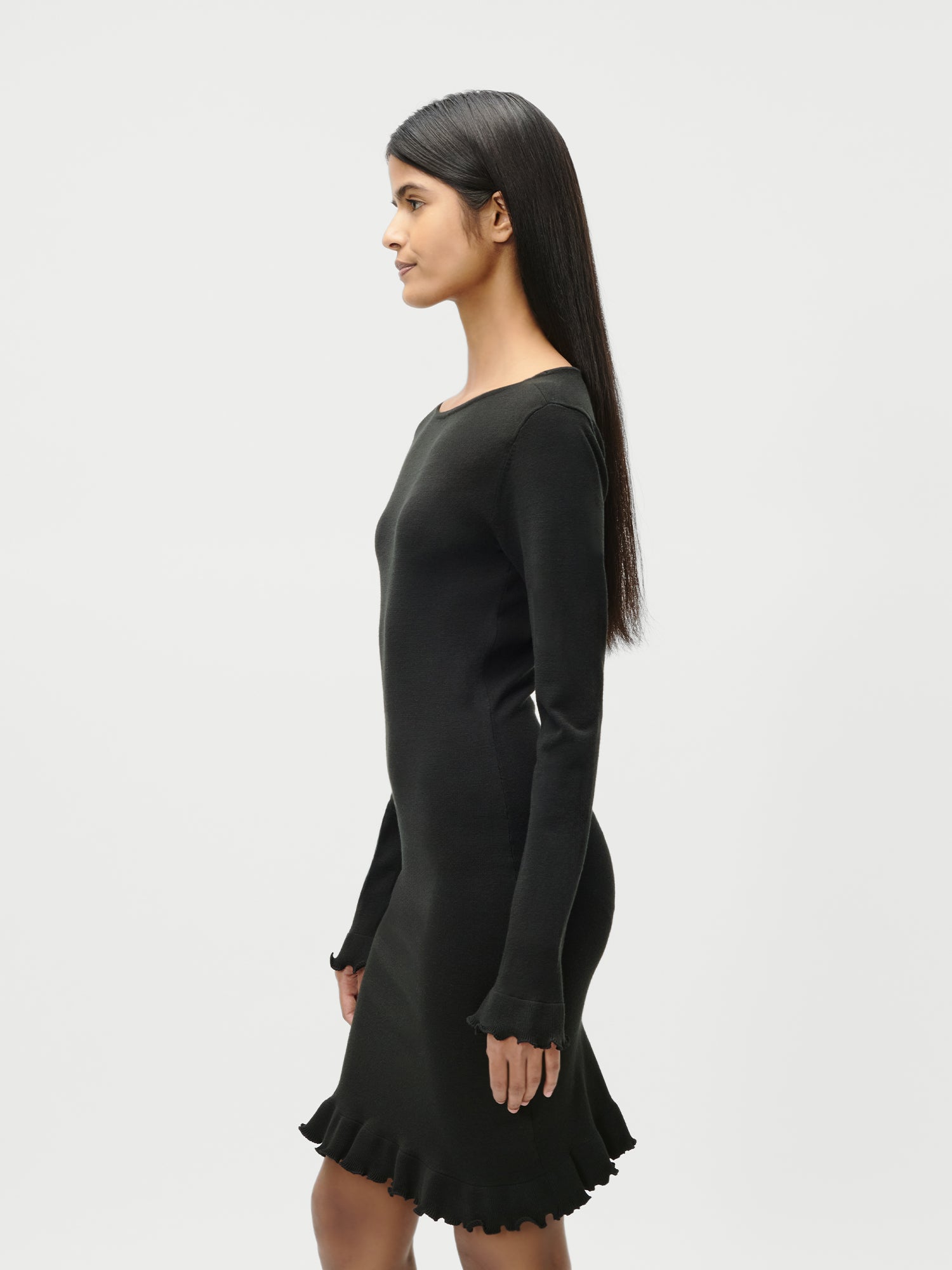 Eine Frau mit langem glattem Haar steht im Profil vor einem schlichten weißen Hintergrund und trägt das LeGer by Lena Gercke Marlen Dress - ein schmal geschnittenes, extralanges schwarzes Kleid mit langen Ärmeln und einem gerüschten Saum.