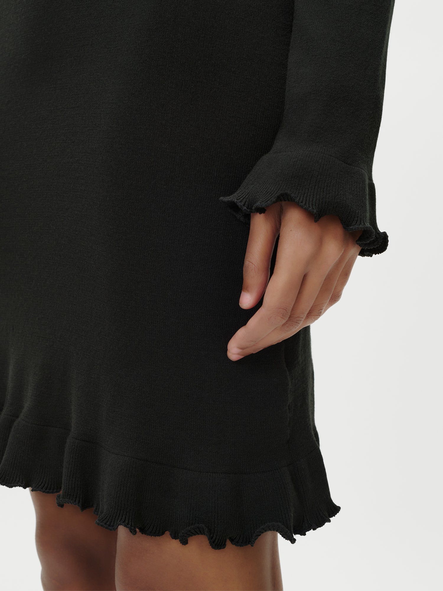 Eine Nahaufnahme einer Person, die das Marlen Dress von LeGer by Lena Gercke trägt - ein schmal geschnittenes, langärmeliges schwarzes Kleid mit gerüschten Manschetten und Saum - zeigt ihre Hand entspannt an ihrer Seite vor einem schlichten weißen Hintergrund.