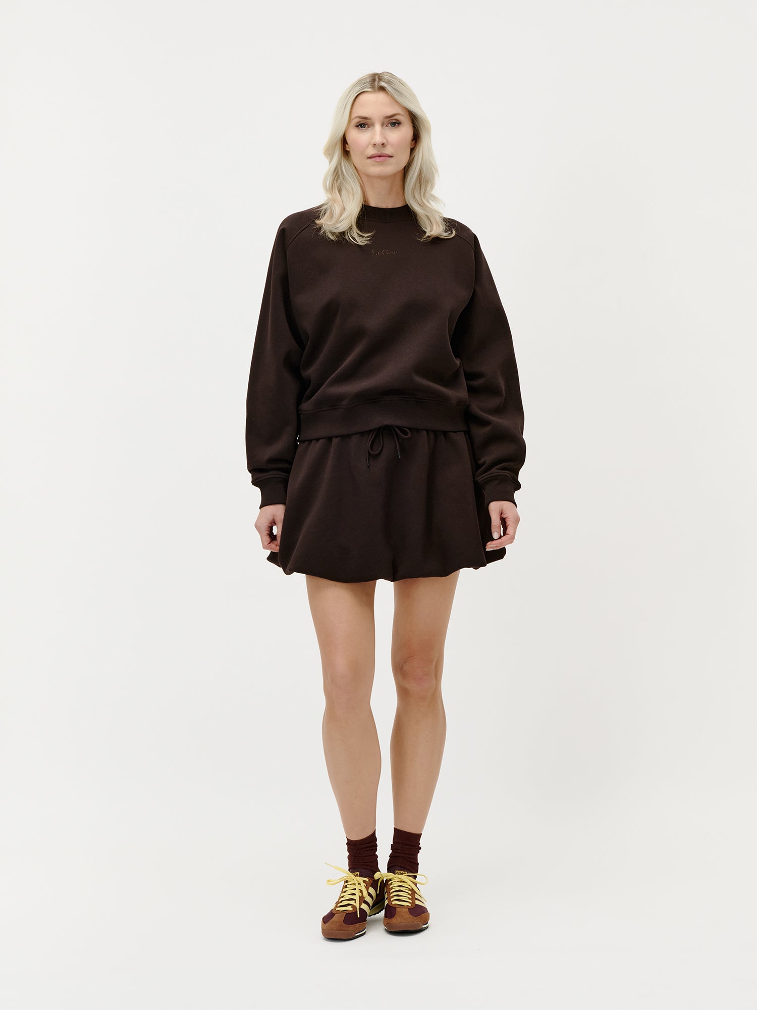 Eine Frau steht vor einem weißen Hintergrund. Sie trägt das lockere dunkelbraune Sweatshirt von LeGer by Lena Gercke im Locker-Style und den dazu passenden Dorina-Rock, dazu kastanienbraune Socken und braune Turnschuhe mit gelben Schnürsenkeln. Sie blickt nach vorne, ihr Gesichtsausdruck ist neutral, ihre Haltung entspannt.