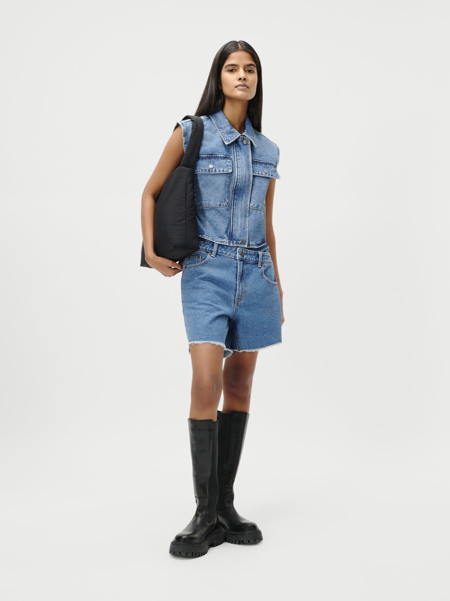 Eine Frau mit langem, glattem Haar und neutralem Gesichtsausdruck steht vor einem weißen Hintergrund, trägt die LeGer by Lena Gercke "Shirin Shorts" Denim-Romper in Größe 36, schwarze, klobige, kniehohe Stiefel und hält eine schwarze, gepolsterte Tasche unter ihrem Arm.
